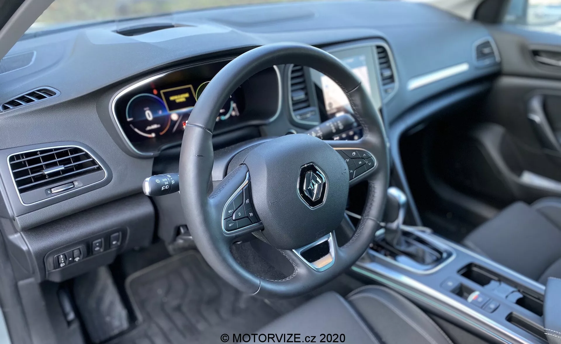 Obrázek zachycuje interiér vozu Renault Megane E-Tech 2022 z pohledu řidiče. Pořízený z mírně odsazeného úhlu zdůrazňuje volant s multifunkčními tlačítky, částečně viditelný digitální přístrojový štít zobrazující barevné ukazatele za ním a infotainmentový systém s tlačítky pod ním v centrální konzoli. Centrální konzole má řadicí páku, což naznačuje automatickou převodovku a různé ovladače. Interiér má kombinaci materiálů včetně kovových lišt a měkkých povrchů. Dobře osvětlený obrázek se ostře zaměřuje na volant a centrální konzoli. V dolní části je vodoznak z MOTORVIZE.cz 2020.
