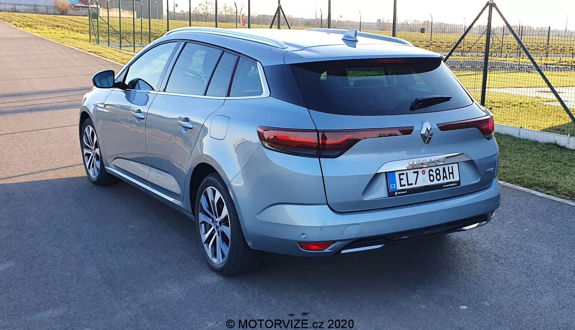 Zadní tři-čtvrté zobrazení hatchbacku Renault Megane-e-Tech 2022, zachycené během dne ze spodní až střední výšky, zaměřené na pravou stranu a záď vozidla. Obsahuje zkosené zadní okno se střešním spoilerem, LED zadní světla, jedinou výfukovou trubku, hliníková kola, zadní stěrač, zadní parkovací kameru a ultrazvukové parkovací senzory. Metalická barva zvyšuje vizuální přitažlivost s evropským stylem na středním umístění registračním čísle, což dále zdůrazňuje tento dobře udržovaný nebo nový vůz.