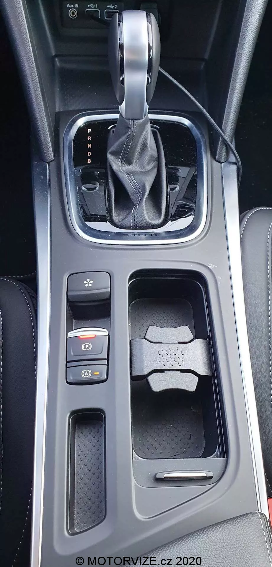 Detailní vertikální záběr na středovou konzolu interiéru vozu Renault Megane E-Tech 2022 z nadhledu. Zobrazuje automatickou převodovku s koženou hlavicí řadicí páky se stříbrným podstavcem v režimu 'park', vysoce lesklý černý rámeček s osvětlenými indikátory PRND, texturovaný povrch za řadicí pákou, tlačítka elektronické parkovací brzdy a Auto Hold, dvojité držáky na nápoje s klíčem v jednom z nich a kombinaci lesklých a texturovaných materiálů s kovovými akcenty.
