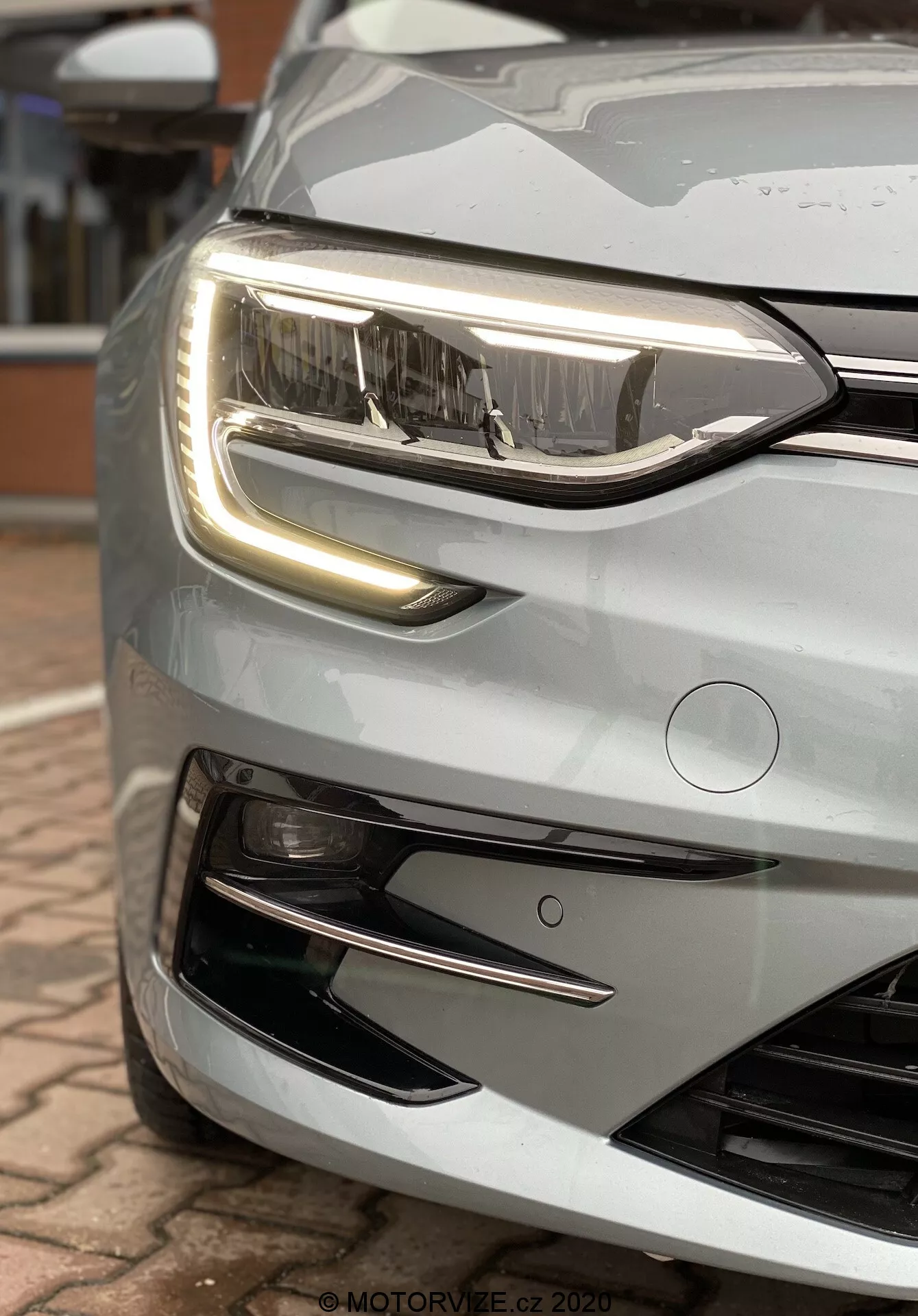 Detailní, šikmý pohled na přední levý díl stříbrného nebo světle šedého vozu Renault Megane E-Tech 2022. Snímek z nízké perspektivy zdůrazňuje přední nárazník, mřížku a světelné prvky. Zaměření je na hlavní světlomet a denní svícení (DRL) s charakteristickým LED designem. Hlavní světlomet má čirou čočku s několika projektorovými světlomety, což naznačuje pokročilou osvětlovací technologii, jako je HID nebo plné LED pro dálková a potkávací světla. DRL má výrazný, nepřerušený světelný pásek, který může fungovat jako ukazatel směru se sekvenčním osvětlováním. Pod DRL má část pro mlhová světla s chromovanou ozdobou, která může hostit přídavná mlhová nebo zatáčecí světla. Dynamický přední nárazník má velkou mřížku přívodu vzduchu s horizontálními lamelami a aerodynamický spodní okraj. Nad mřížkou se nachází kruhový senzor, naznačující pokročilé asistenční systémy řidiče (ADAS). Kapota klesá dolů s měkkými obrysy, ukazuje elegantní a čistý přední profil, navzdory kapkám deště naznačujícímu venkovní prostředí. Obrázek ukazuje moderní automobilový design a pokročilé technologické prvky.