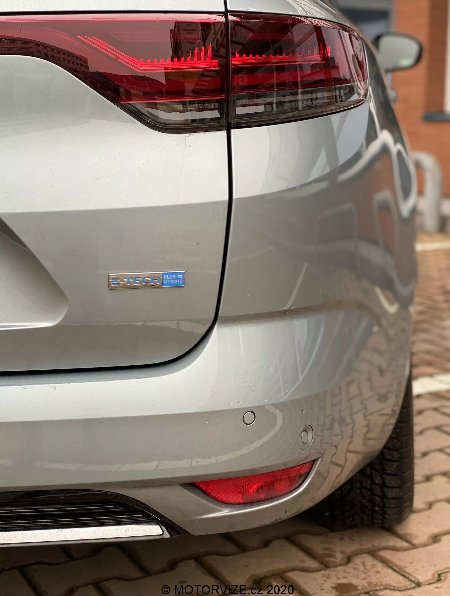 Blízký pohled na zadní roh moderního vozu Renault Megane 2022 (Megane-E-Tech) z nízkého a zadního úhlu. Obrázek zdůrazňuje červené a průhledné zadní světlo, které se rozšiřuje do elegantního zadního čtvrtletního panelu, jemně zakřivené víko zavazadlového prostoru s rozmazaným odznakem pro větší soukromí naznačující hybridní nebo elektrickou variantu. Mezi viditelné vlastnosti patří kruhové parkovací senzory pod zadním světlem, lesklá kovová koncovka výfuku integrovaná do zakřiveného zadního nárazníku a kovová stříbrná reflexní barva zachytávající okolní objekty.
