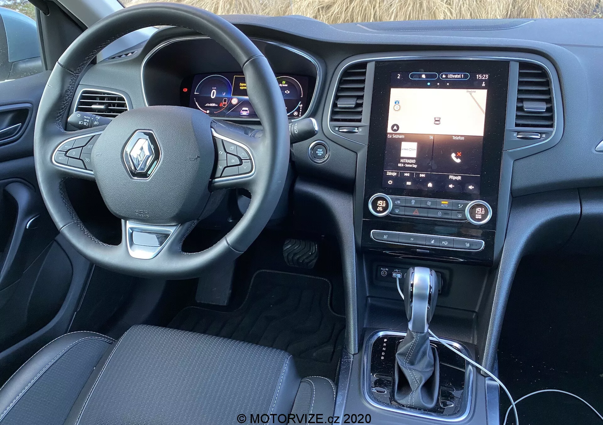 Interiér vozu Renault Megane 2022 z pohledu řidiče, zobrazující volant s logem Renault, digitální přístrojovou desku a vertikálně orientovaný dotykový infotainment systém na středové konzoli. Podrobný pohled zahrnuje tlačítka pro různé funkce na volantu, osvícené ukazatele vozidla, řadicí páku automatické převodovky a minimální fyzické ovladače pod infotainment systémem.