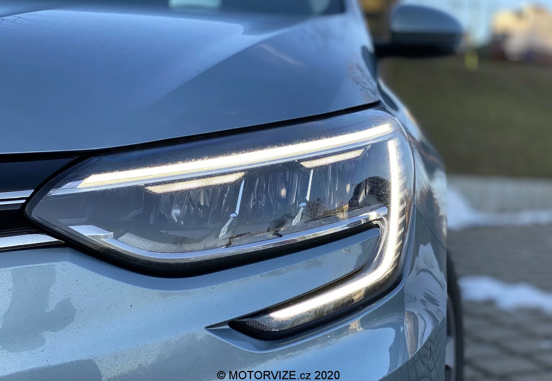 Detailní záběr přední části vozu Renault Megane-e-Tech 2022, zachycující složitě navržený LED světlomet s úhlovým denním světlem a částečně viditelnou lamelovou přední mřížkou. Obrázek se zaměřuje na lehce zvýšený boční úhel a zvýrazňuje aerodynamické linie karoserie, lesklý kovový exteriér a pokročilou osvětlovací technologii s profesionálním efektem bokeh v pozadí.