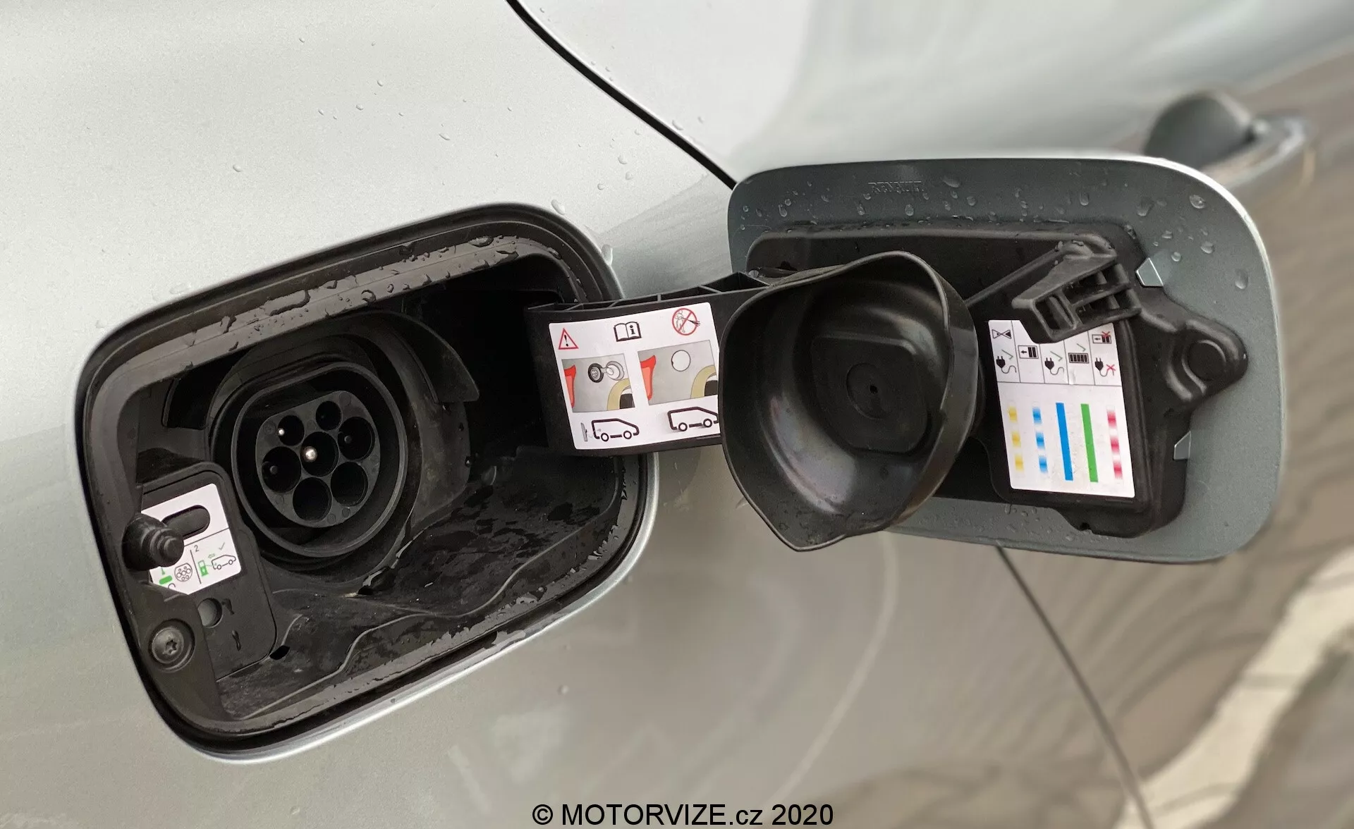 Detailní pohled na oblast nabíjecího portu elektromobilu (EV) z diagonálního úhlu se zaměřením na otevřená dvířka nabíjecího portu, která odhalují konfiguraci zásuvek. Viditelná je karoserie vozidla obklopující prostor víčka nádrže, což naznačuje, že se jedná o světlé vozidlo. Zobrazeny jsou dvě samostatné zásuvky, které naznačují podporu jak AC, tak DC nabíjení, jedna se podobá Type 2 konektoru pro AC a druhá odpovídá CCS konektoru pro rychlé nabíjení DC. Označení kolem portů obsahuje symboly pro typy nabíjení, informace o napětí a samolepku s QR kódem a barevnými pruhy. Dvířka nabíjecího portu jsou pružinově zatížená se zřetelnými nečistotami a vodou, což naznačuje pravidelné používání a vystavení vlivům počasí. Obrázek je pořízen mírně směrem dolů k zásuvkám.