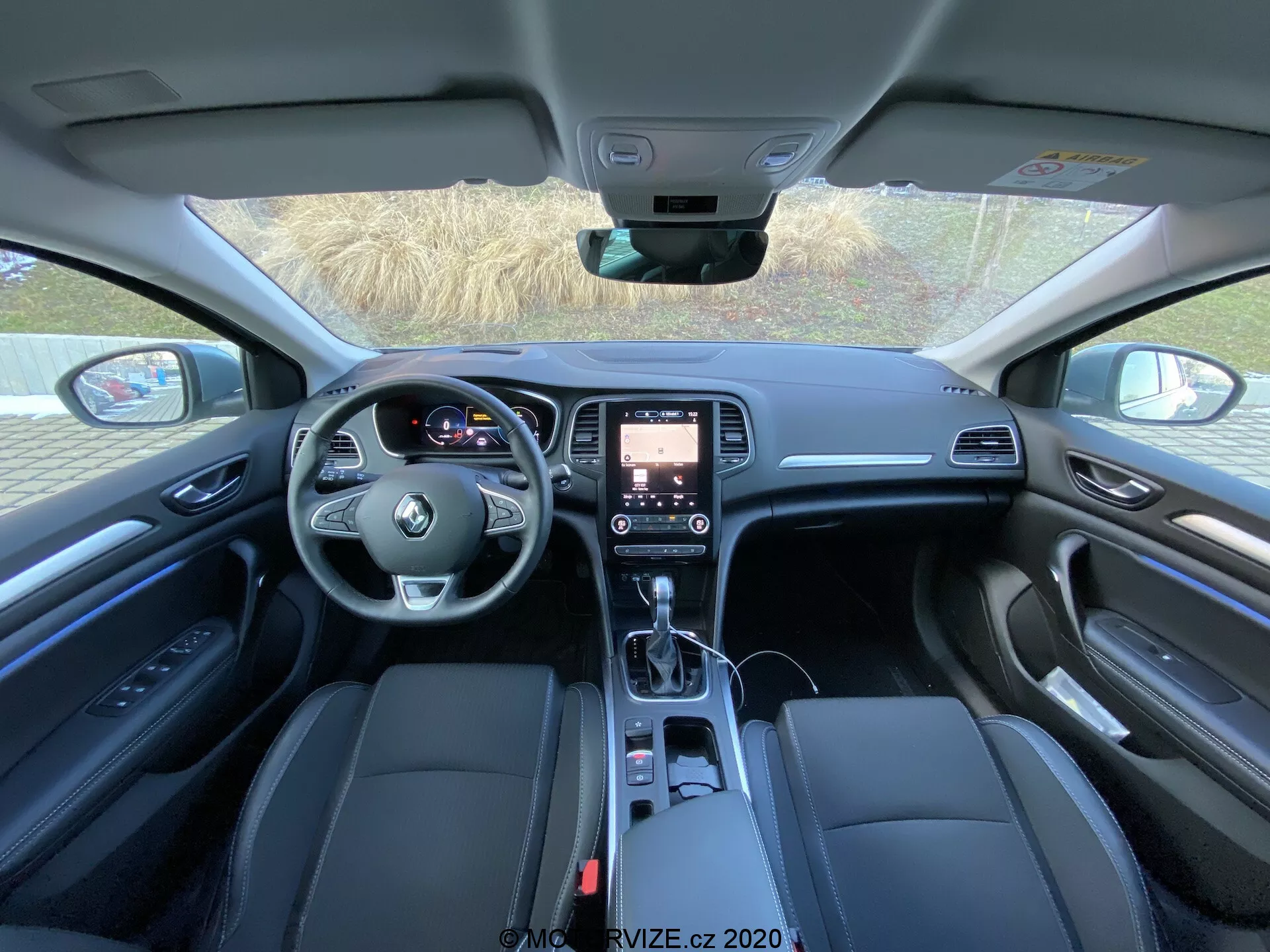 Pohled na interiér Renault Megane E-Tech 2022 ze zadního sedadla, zobrazující moderní palubní desku s ovládacími prvky na levostranném volantu, dotykovou infotainment obrazovku, automatickou řadicí páku, středovou konzoli s držáky na nápoje a úložištěm, kožená čalouněná přední sedadla s elektronickým nastavením, dveřní panely s paměťovým nastavením a reproduktorem audiosystému, a kvalitní materiály obložení.