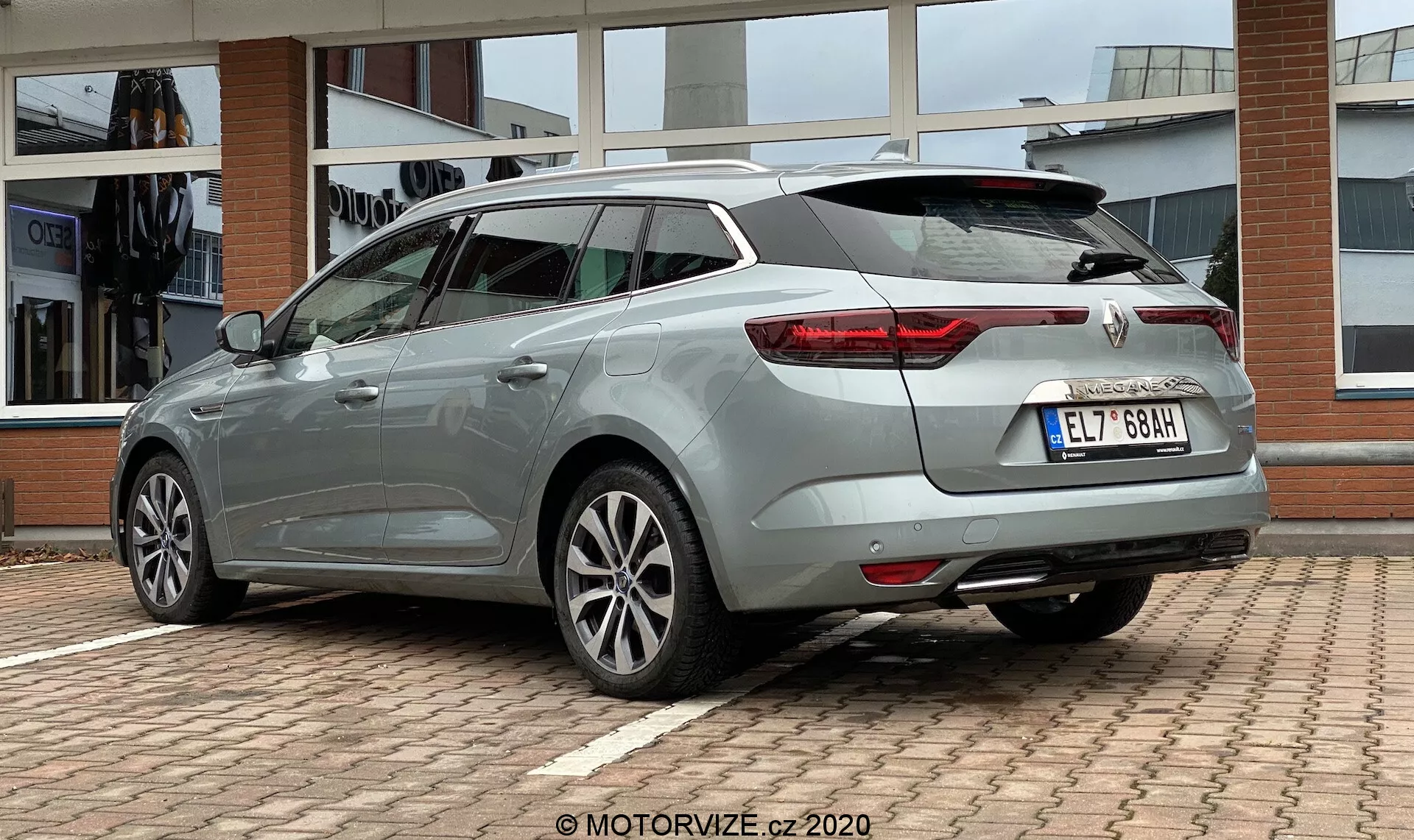 Tříčtvrteční pohled zezadu na Renault Megane E-Tech 2022, zdůrazňující jeho prodlouženou střechu a zadní část. Obrázek ukazuje výrazná LED zadní světla, stříbrná vícepaprsková kola, boční prahy, zadní stěrač, vysoko umístěné brzdové světlo integrované do zadního spoileru a žraločí anténu. Auto má světlý metalický modro-šedý lesklý lak s označením značky a modelu na zadní straně. Vozidlo je umístěno na parkovišti s fasádou budovy v pozadí, snímáno z nízkého úhlu pro zdůraznění jeho designu.