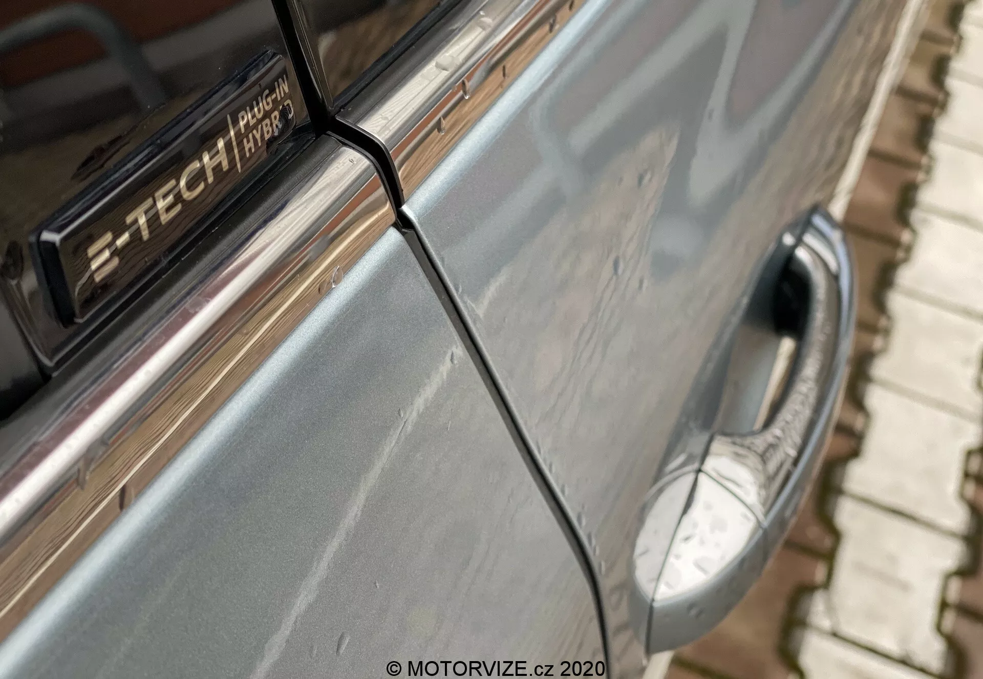 Detailní pohled na Renault Megane 2022 (Megane-e-Tech), zaměřený na stranu vozu těsně za předním blatníkem, kde se setkávají přední dveře s těmi zadními. Vystažen je ostrý svislý šev, chromovaná klika dveří, lesklý šedý lak s drobnými nedokonalostmi a odznak 'TECH'. V rozmazaném popředí je viditelné boční zrcátko.