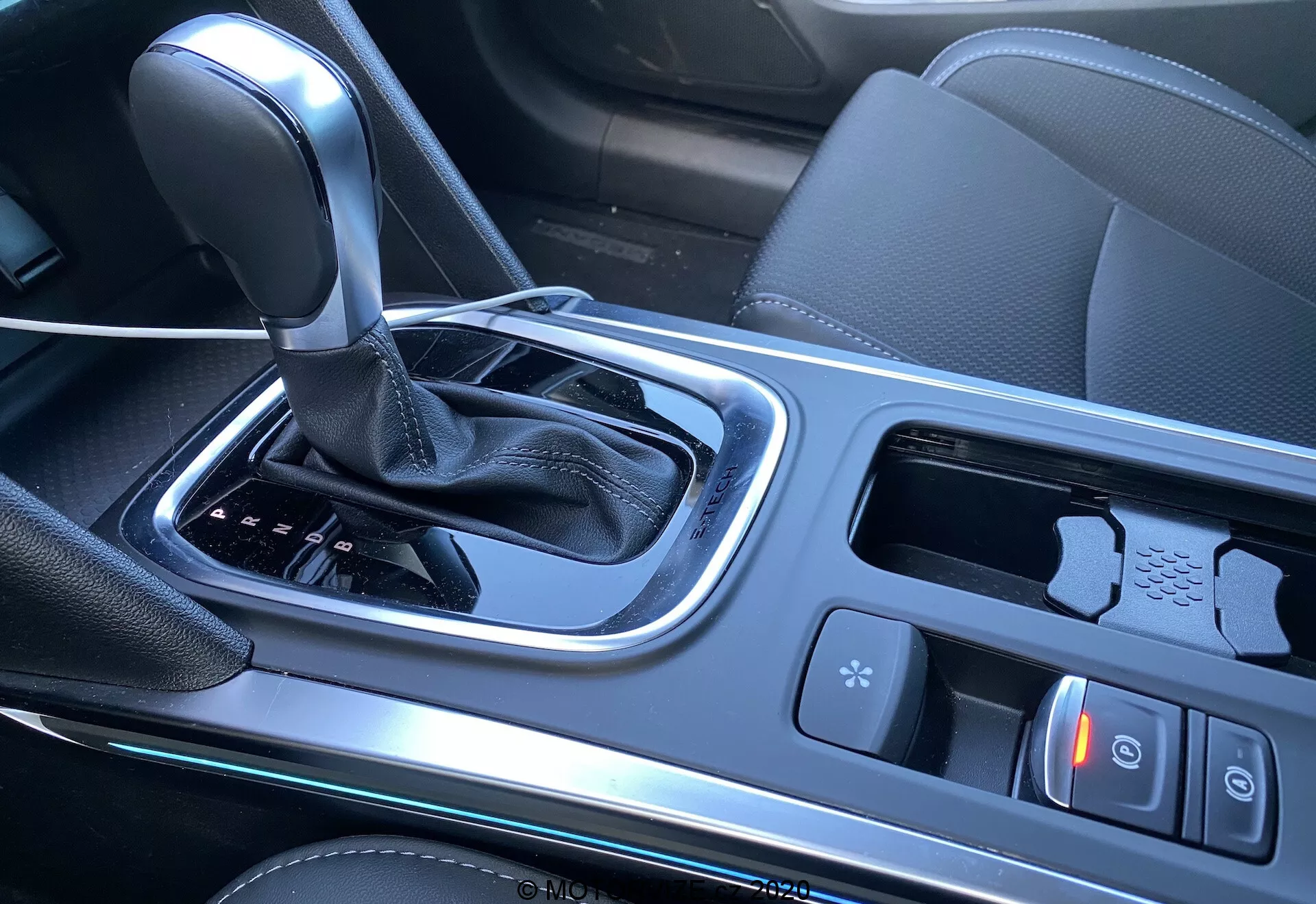 Renault Megane E-Tech 2022, středová konzole interiéru se zaměřením na oblast řadicí páky, zahrnující automatickou převodovku s koženou rukojetí, ovladače jízdních režimů, tlačítko elektrické parkovací brzdy, držáky na nápoje, dotykový panel nebo joystick pro multimediální/navigační systémy, malý úložný prostor pravděpodobně s USB porty a perforovaný kožený okraj sedadla. Leskle černé plastové panely a kovové lišty, naznačující moderní a technologicky orientované auto.