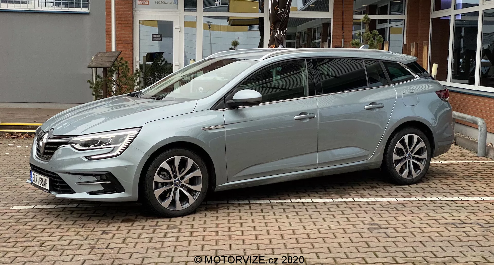Boční profil Renaultu Megane 2022 (Megane-e-Tech) zaparkovaného na cihlové dlažbě. Obrázek ukazuje pětidveřový hatchback s litinovými koly, zpětnými zrcátky s integrovanými směrovkami a elegantním stříbrným nebo světle šedým povrchem. Auto má ostré linie podél klik dveří a klesající střechu. Viditelný chrom v rámech oken přidává eleganci. Vozidlo je čisté a stabilní, nakloněno tak, aby ukazovalo tři čtvrtiny pohledu na stranu spolujezdce, pod zataženým nebem.