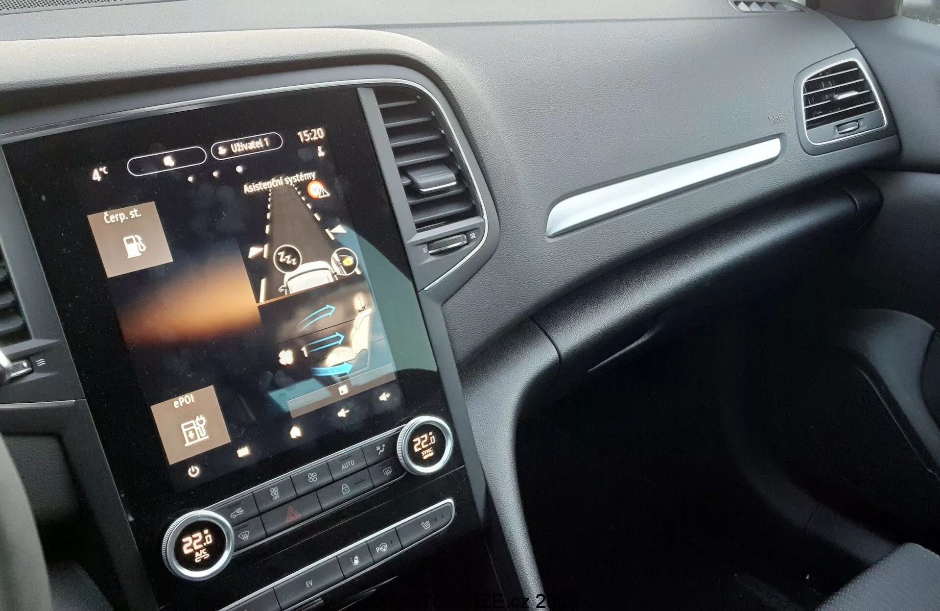 Detailní pohled na středovou konzoli interiéru vozu Renault Megane 2022 (Megane-e-Tech), zdůrazňující velký vertikální dotykový displej infotainment systému s ikonami pro přehrávání médií, nastavení vozidla, klimatizaci a navigaci. Pod displejem jsou fyzické tlačítka a ovladače systému HVAC, dvouzónová klimatizace nastavená na 22 stupňů Celsia. Tmavá palubní deska s kontrastní povrchovou úpravou z kartáčovaného kovu a větracími otvory nad displejem. Částečný pohled na volant z úhlu, snímek pořízený z předního sedadla spolujezdce.