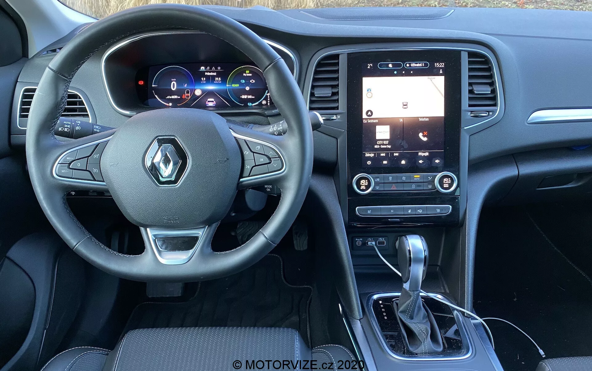Pohled na interiér Renaultu Megane-E-Tech 2022 z perspektivy spolujezdce, zobrazující volant s logem Renault a ovládací prvky, digitální přístrojový panel zobrazující informace o vozidle, vertikálně orientovaný dotykový displej na středové konzoli s funkcemi infotainmentu jako GPS navigace a nastavení zvuku, automatickou převodovku s koženým potahem, držáky na nápoje, vertikální větrací otvory s nastavením klimatizace a prémiové interiérové materiály včetně tmavých plastů a kartáčovaného kovu kolem řadicí páky a volantu, s viditelnými okraji kožených nebo syntetických sedadel.