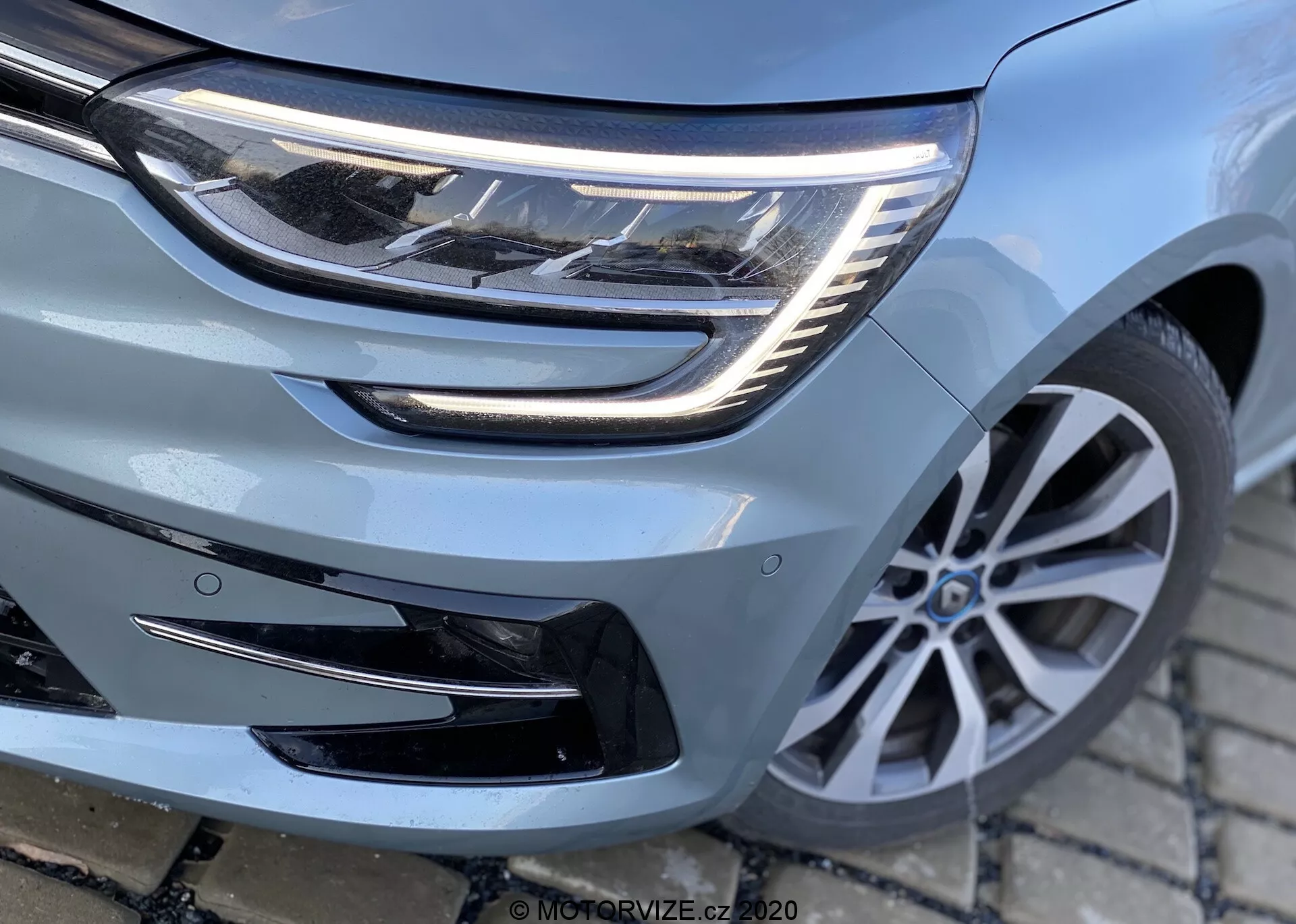 Detailní pohled zepředu na Renault Megane 2022 Megane-e-Tech se zaměřením na světlomet, mřížku a přední nárazník, s ukázkou LED světelných prvků, horizontálního designu mřížky a potenciálního slotu pro senzory asistence řidiče. Díky mnohapaprskovému designu kol a modernímu stylu vozidla z nízkého úhlu, mírně vlevo.