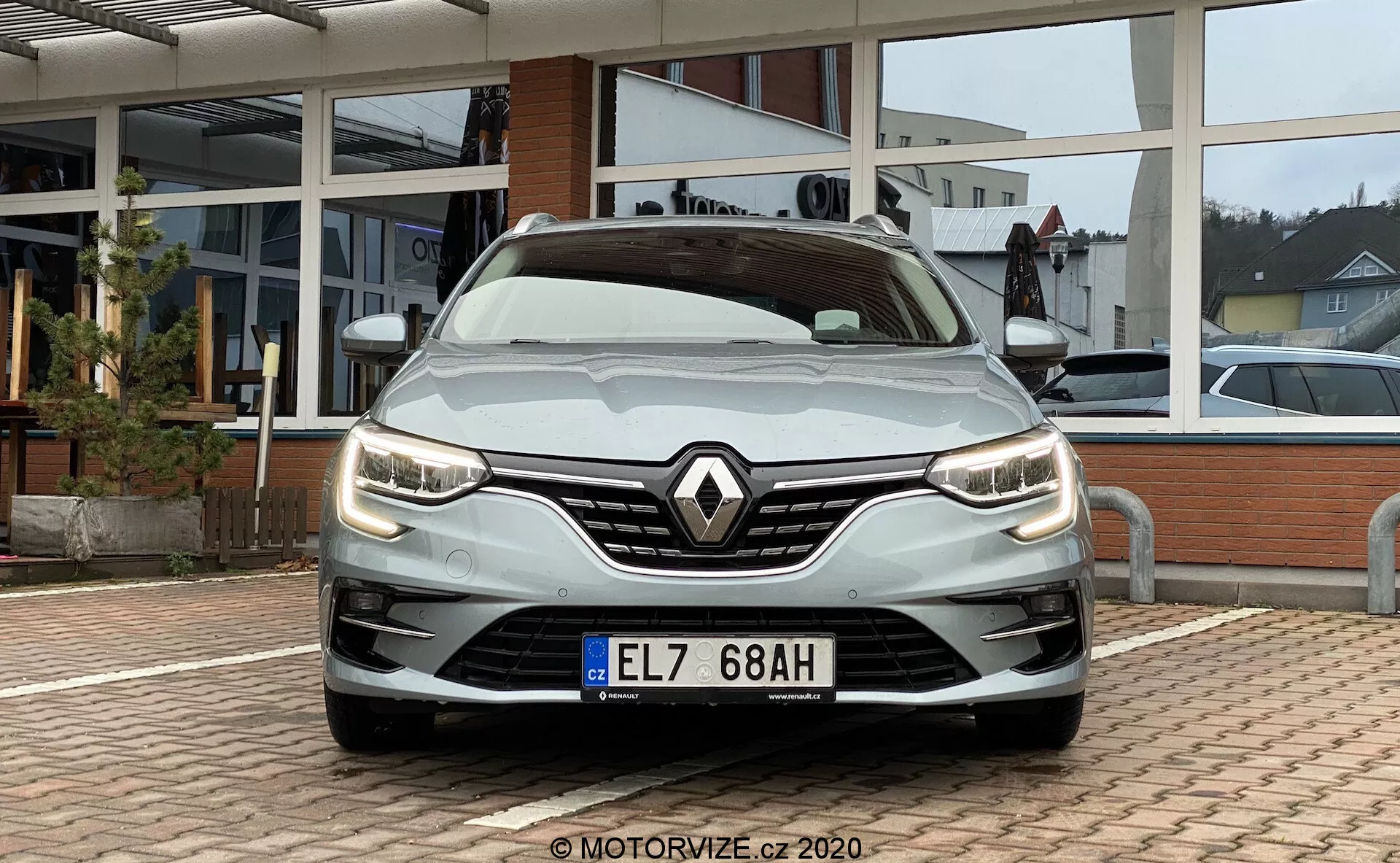 Pohled zepředu na stříbrný Renault Megane E-Tech z roku 2022 z nízkého úhlu blízko mřížky chladiče. Fotografie zachycuje stylové moderní LED světlomety, výrazný znak Renault na mřížce s kombinovaným vzorem a aerodynamické obrysy kapoty. Přední nárazník integruje mlhovky a přívody vzduchu, zdůrazňující estetiku a funkčnost. Přírodní denní světlo s oblačnými podmínkami osvětluje vozidlo, s dlážděným prostorem a prosklenou fasádou autosalonu v pozadí.