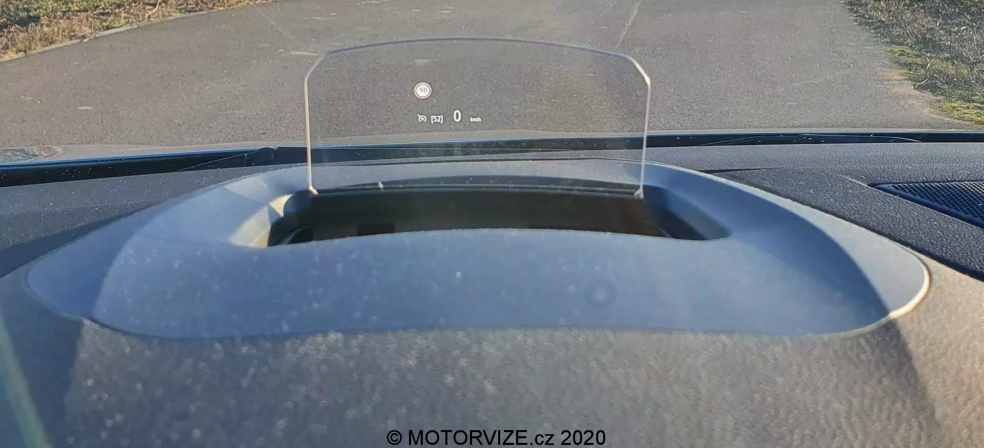 Interiér z pohledu řidiče Renaultu Megane E-Tech 2022, zaměřující se na head-up display promítající klíčové jízdní údaje jako rychlost a navigaci na průhlednou obrazovku nad palubní deskou. Přední sklo ukazuje jednoproudou silnici s vegetací po obou stranách, což naznačuje předměstské nebo venkovské prostředí. Matný povrch kapoty a viditelné částečky prachu naznačují nedávné použití nebo potřebu čištění.