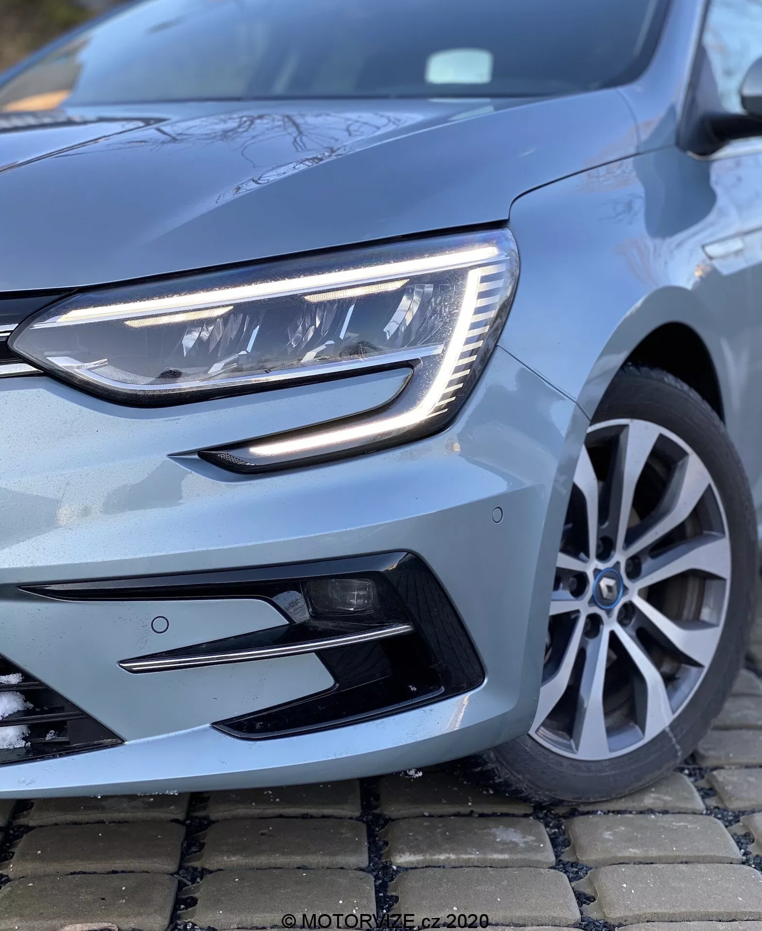 Detailní pohled na Renault Megane 2022 (Megane-e-Tech) z nízkého úhlu, zaměřený na levý přední čtvrtinový pohled. Obraz výrazně zobrazuje moderní světlomet s LED prvky a elegantní denní světlo. Je vidět část přední mřížky a nárazníku s černými akcentovanými přívody vzduchu, spolu se stylovým vícespeicovým slitinovým kolem a kovově světle modrým lakem, který odráží péči a stav vozu.
