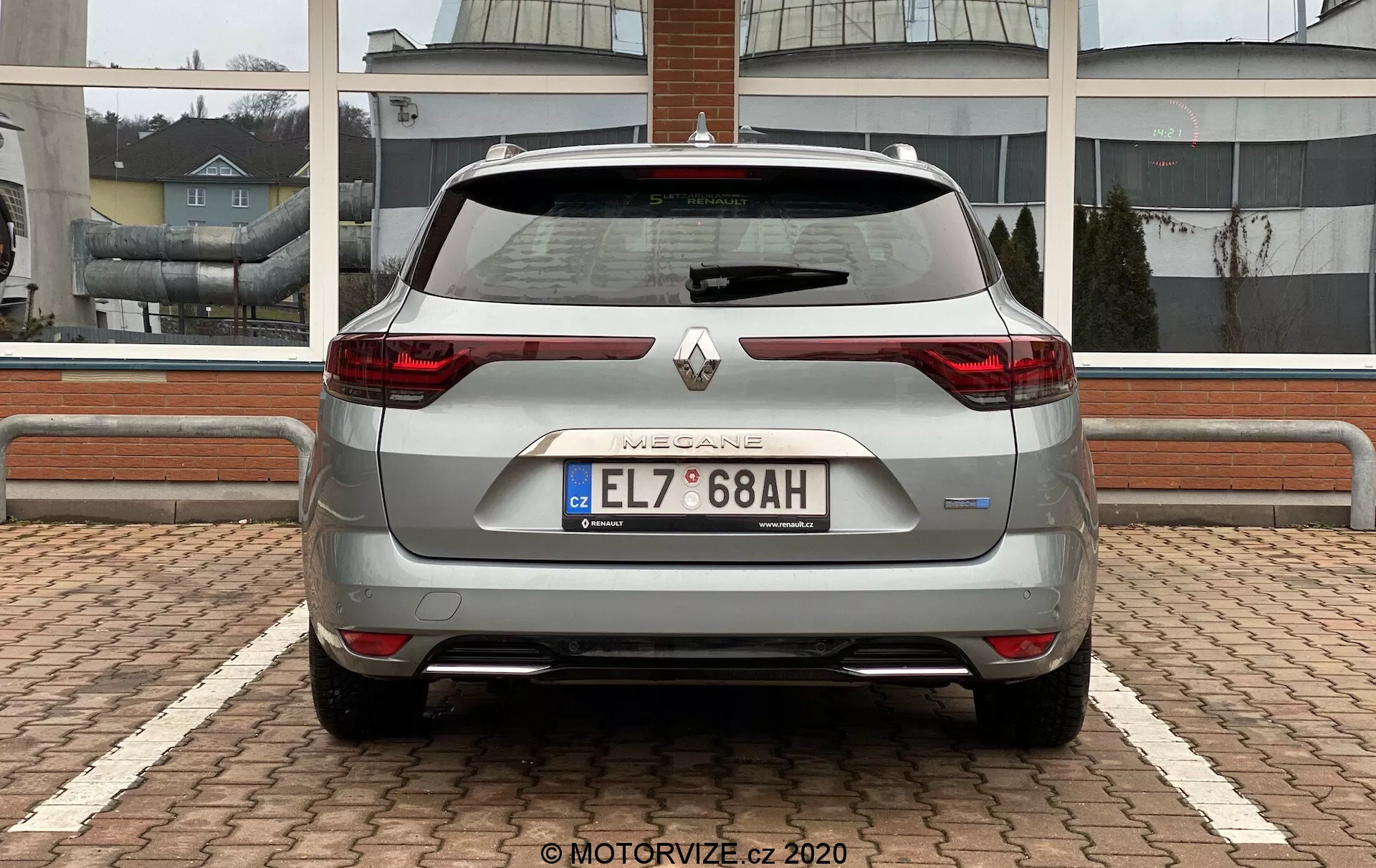 Pohled zezadu na stříbrný Renault Megane E-Tech 2022 s LED zadními světly ve tvaru C, středovým logem Renault pod nápisem 'MEGANE', evropskou poznávací značkou, parkovacími senzory, vnějšími odrazkami, zadním stěračem na zadním okně, mírně klesající linií střechy končící střešním spoilerem, anténou ve tvaru žraločí ploutve, čistým stylistickým vzhledem zadní části s ukrytým výfukem a částečně viditelnými hliníkovými koly. Vozidlo je zaparkováno na dlažbě ve vzoru rybí kosti, na pozadí je obchodní nebo obytná budova.