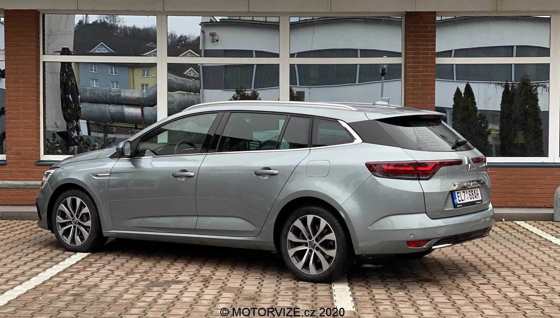 Pohled z profilu na stříbrně šedý Renaut Megane 2022 kombi, zaparkovaný na povrchu s cihlovým vzorem se štíhlými obrysovými liniemi, zrcátky a klikami dveří v barvě karoserie, vícepaprskovými litými koly, výraznými obtočnými zadními světly, zadním stěračem, vysoko umístěným brzdovým světlem a střešním spoilerem. Auto je umístěno proti budově s velkými okny, které odrážejí zataženou oblohu.