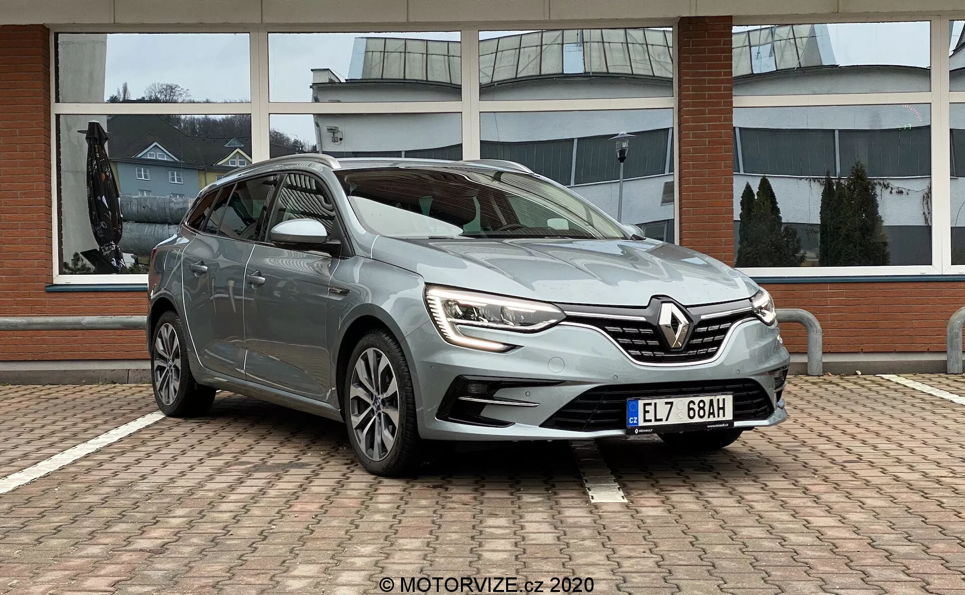 Pohled z předního tříčtvrtního úhlu na hatchback Renault Megane 2022 (Megane E-Tech), zaparkovaný na dlážděné ploše s komerční budovou s velkými skleněnými okny v pozadí. Záběr z předního úhlu řidičovy strany zvýrazňuje LED denní svícení, propracovaný design mřížky s logem Renault, vytvarované boční linie, vícelúčková litá kola a boční zrcátka s integrovanými směrovkami. Pochmurné počasí poskytuje měkké, rozptýlené osvětlení, které zvyšuje viditelnost designových prvků.