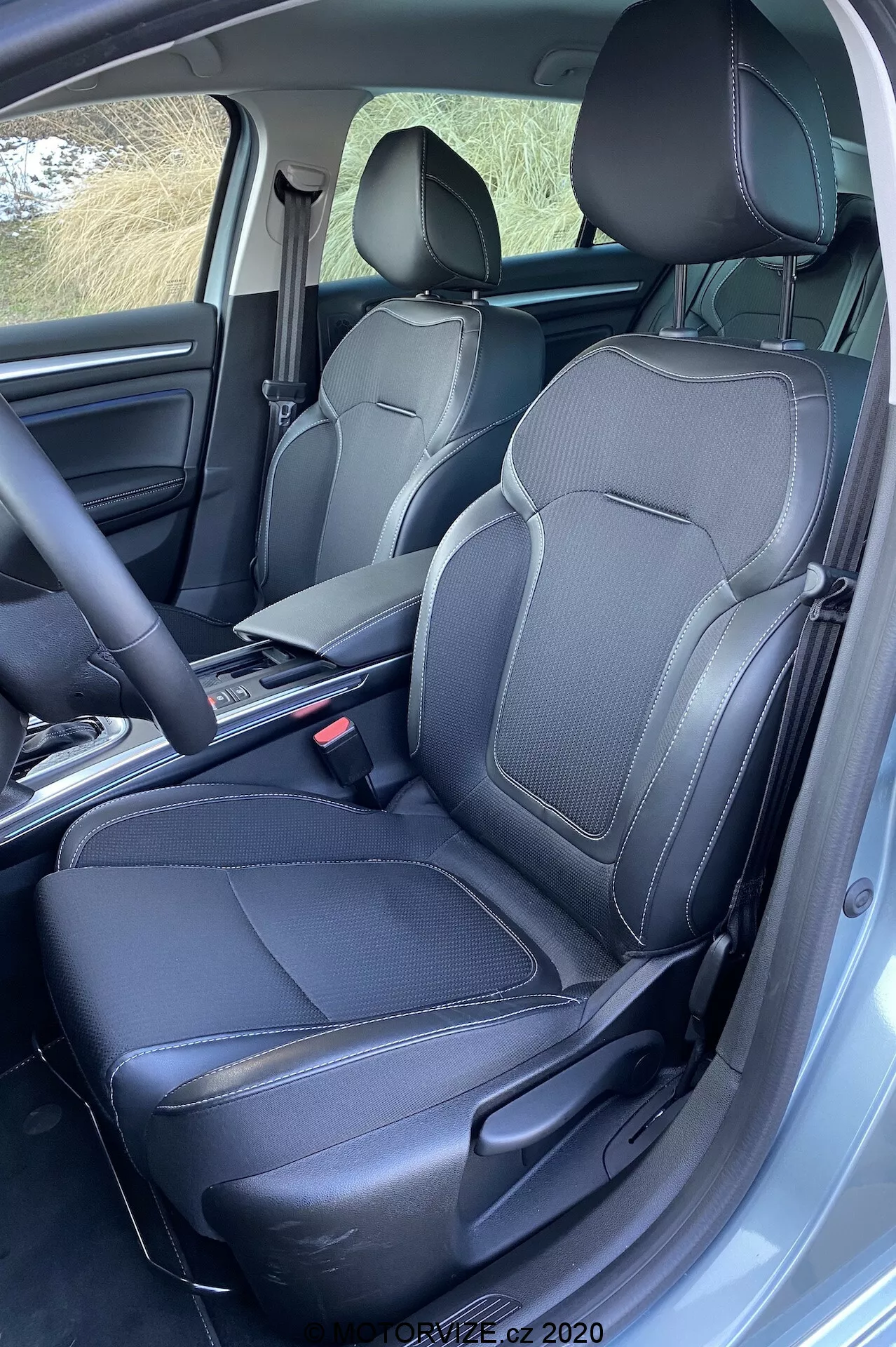 Renault Megane 2022 interiér přední prostor pro spolujezdce s černými koženými sedadly s detailním švem, opěrkami hlavy a boční oporou. Středová konzola s nejasnou řadicí pákou a sklopenou loketní opěrkou, poskytující úložný prostor. Viditelné bezpečnostní pásy a mechanismus kotvení, koberce s rohožemi na míru. Otevřené dveře na straně řidiče, mírně zvýšený úhel pohledu ukazující prvotřídní provedení.