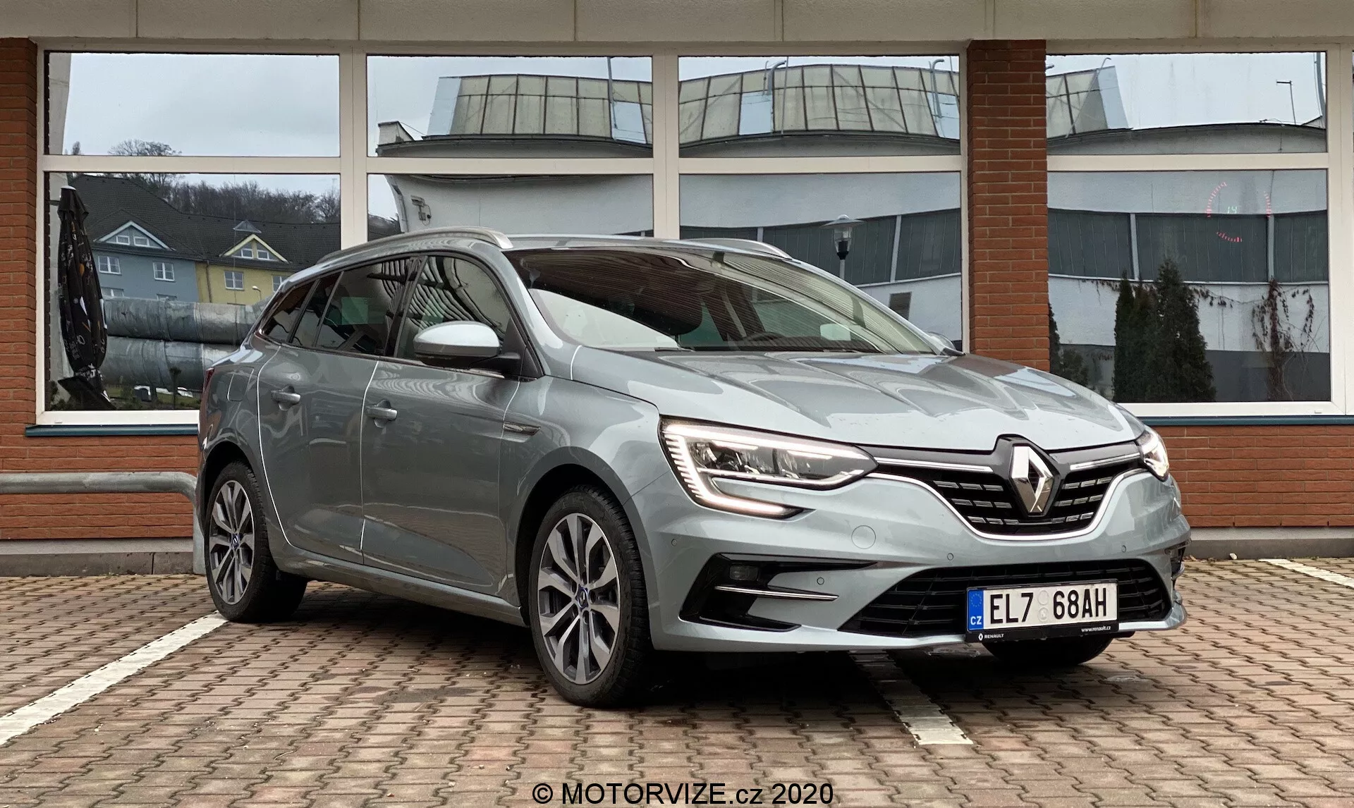 Stále stojící Renault Megane E-Tech 2022 zobrazený z pohledu tři čtvrtiny spredu, zdůrazňující elegantní přední mřížku s integrovanými LED denními světly, mlhovky a aerodynamický nárazník. Boční profil ukazuje panely dveří řidiče, okna, zrcátka s blikajícími světly a víceramenná litá kola. Metalický lak, lesklý povrch, zaparkovaný na vzorované cihlové ploše, pravděpodobně u prodejce. Přirozené osvětlení zlepšuje čistý, dobře udržovaný vzhled.