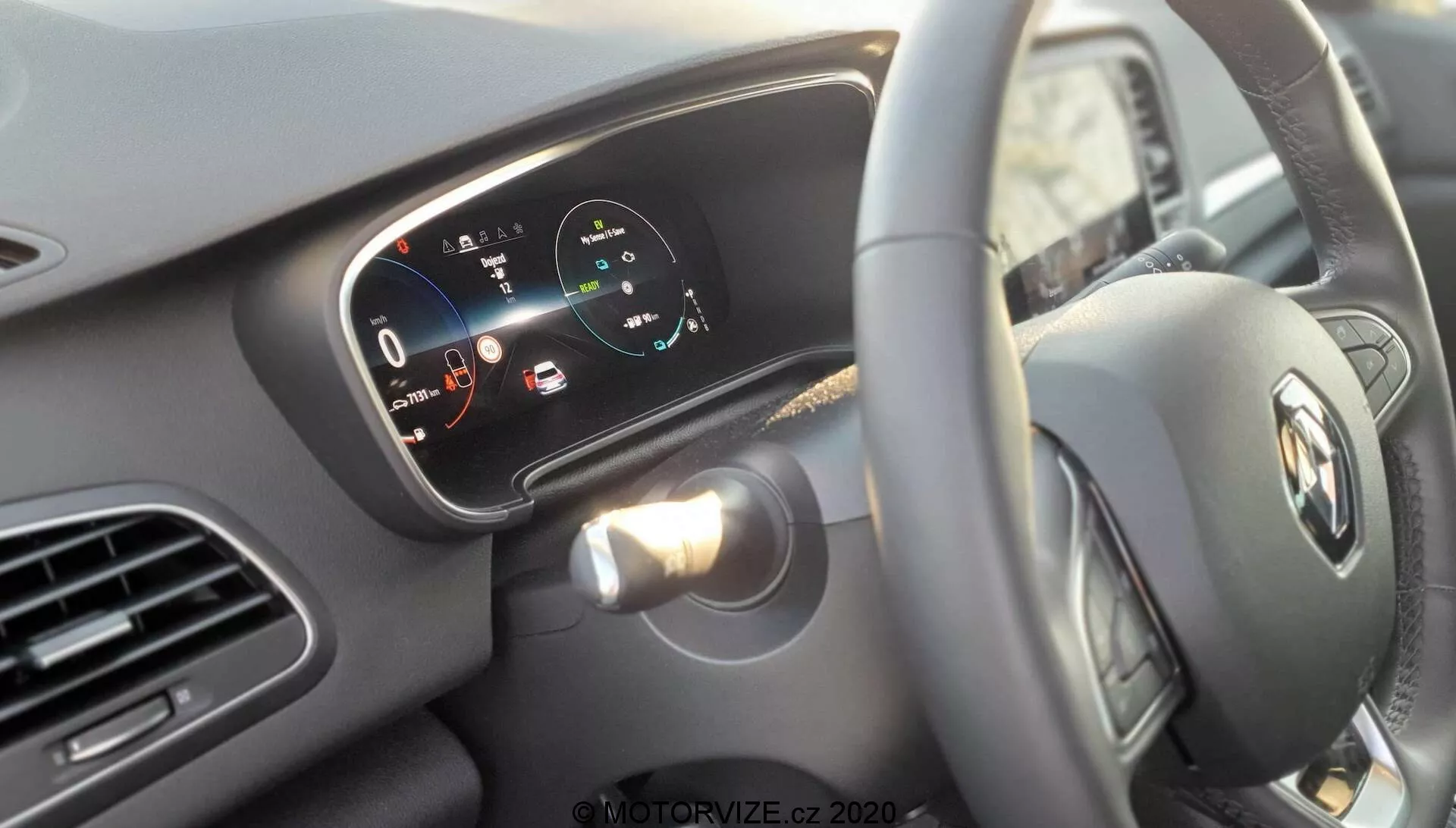 Pohled na interiér vozu Renault Megane E-Tech 2022 z pohledu řidiče, zaměřený na volant s logem Renault a digitální přístrojovou desku za ním. Sloupek řízení má vlevo jednu páčku pro směrovky a možná i ovládání světel. Digitální displej zobrazuje rychloměr s hodnotou 0 km/h a ukazatel výkonu s označením „charge“, „eco“ a „power“, naznačující elektrickou nebo hybridní povahu vozidla. Jsou vidět výstražná světla a ikony pro úroveň nabití baterie, otevřené dveře a další stavy. Vleva je vidět část infotainmentu nebo navigačního systému. Minimalistický interiérový design s horizontálními liniemi přes palubní desku a ventilátory zdůrazňuje šířku vozidla. Ostření zdůrazňuje emblém volantu a digitální displej, čímž podtrhuje pokročilé digitální rozhraní palubní desky.