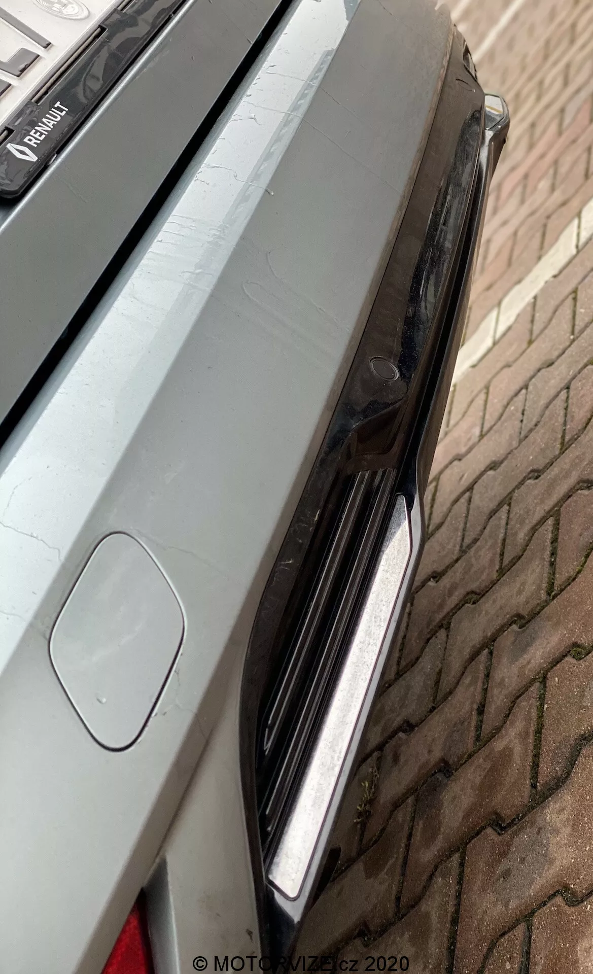 Detailní pohled na zadní pravou část vozu Renault Megane E-Tech z roku 2022, se zaměřením na zadní světelnou sestavu s LED světly, okraj víka kufru s integrovaným spoilerem a malou část zadního nárazníku s možným parkovacím senzorem nebo zadní kamerou. Vůz má čistý metalický lak, který odráží jeho okolí, a je umístěn na cihlové dlažbě.
