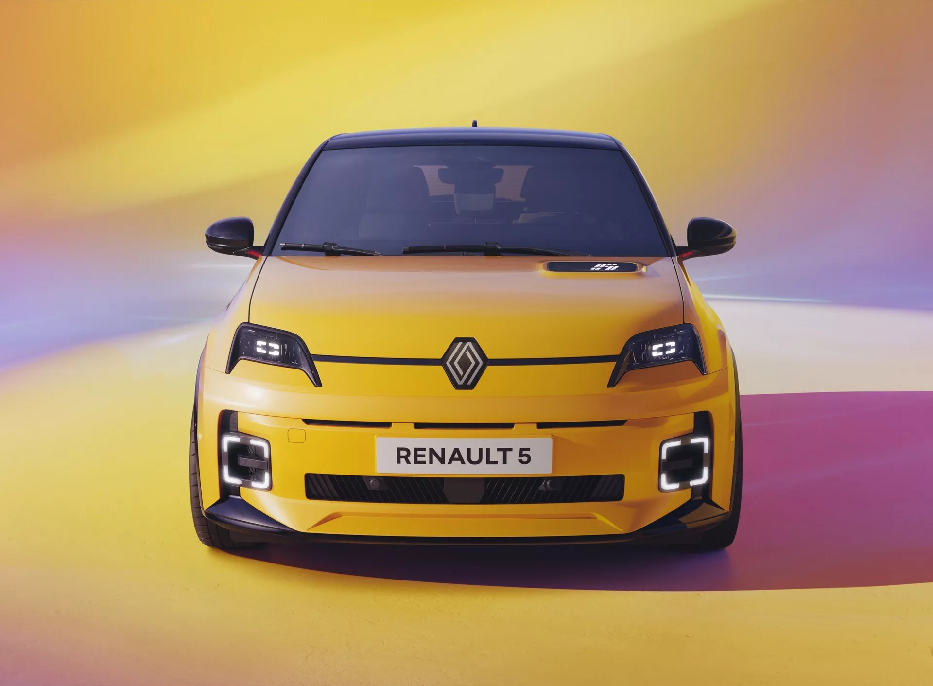Živý pohled zepředu na Renault 5 ročník 2024 v jasně žluté barvě na pozadí s přechodem od fialové k žluté, zvýrazňující jeho moderní LED světlomety, velké nasávací otvory na nárazníku a sportovní design.