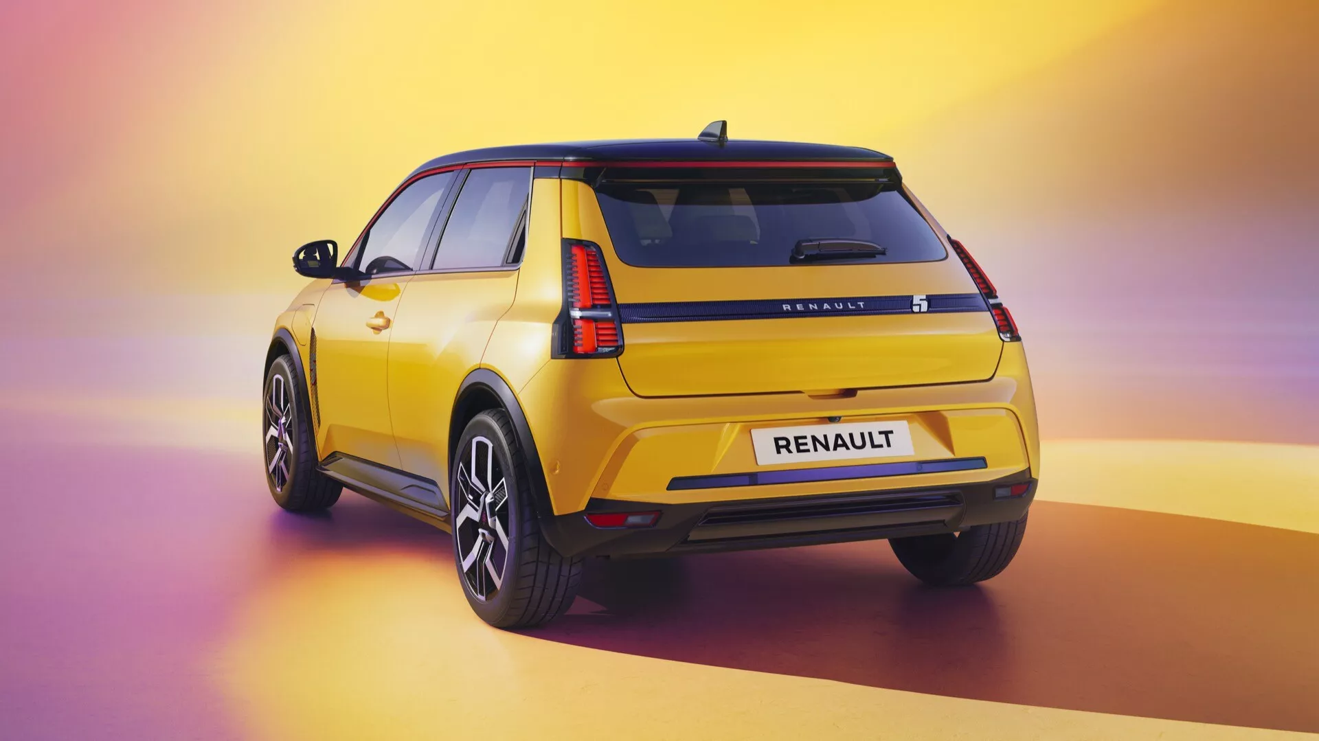 Tříčtvrteční zadní pohled na žlutý Renault R5 (III) rok 2024 ukazuje LED zadní světla, značení RENAULT, zadní okno se stěračem, litá kola s více paprsky, černé lišty, spoiler na střeše, leštěný lak a anténu ve tvaru žraločí ploutve na pozadí s přechodem