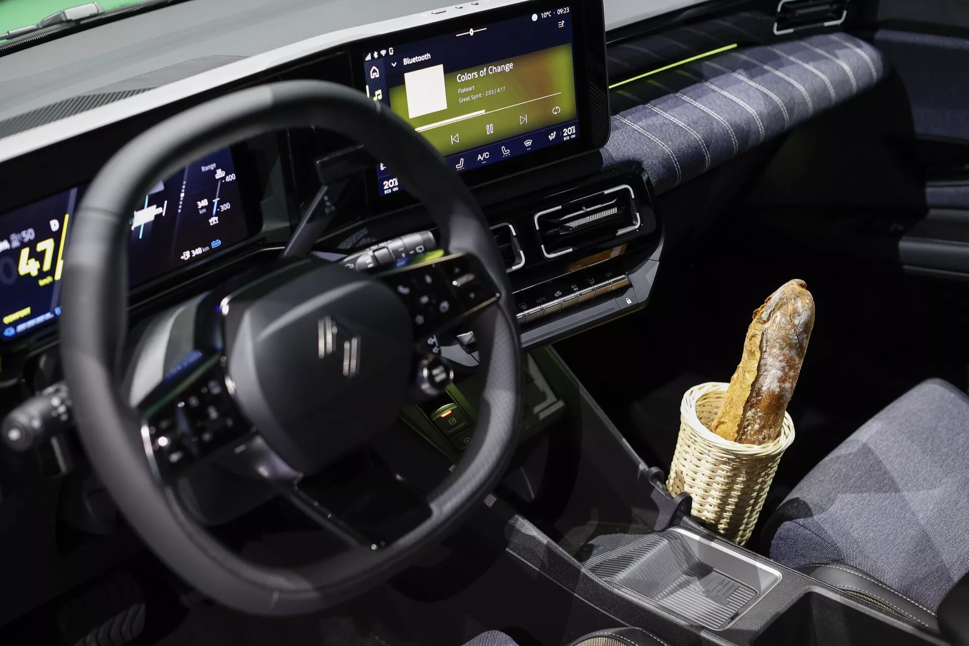 Interiér Renaultu R5 2024 prezentující multifunkční volant s plochým dnem, široký digitální přístrojový panel zobrazující stav elektrického vozidla jako rychlost a stav baterie, centrální dotykový displej informačního systému s možnostmi připojení a vtipný prvek v podobě bagety ve středovém úložném prostoru, odrážející moderní design a technologie vozidla