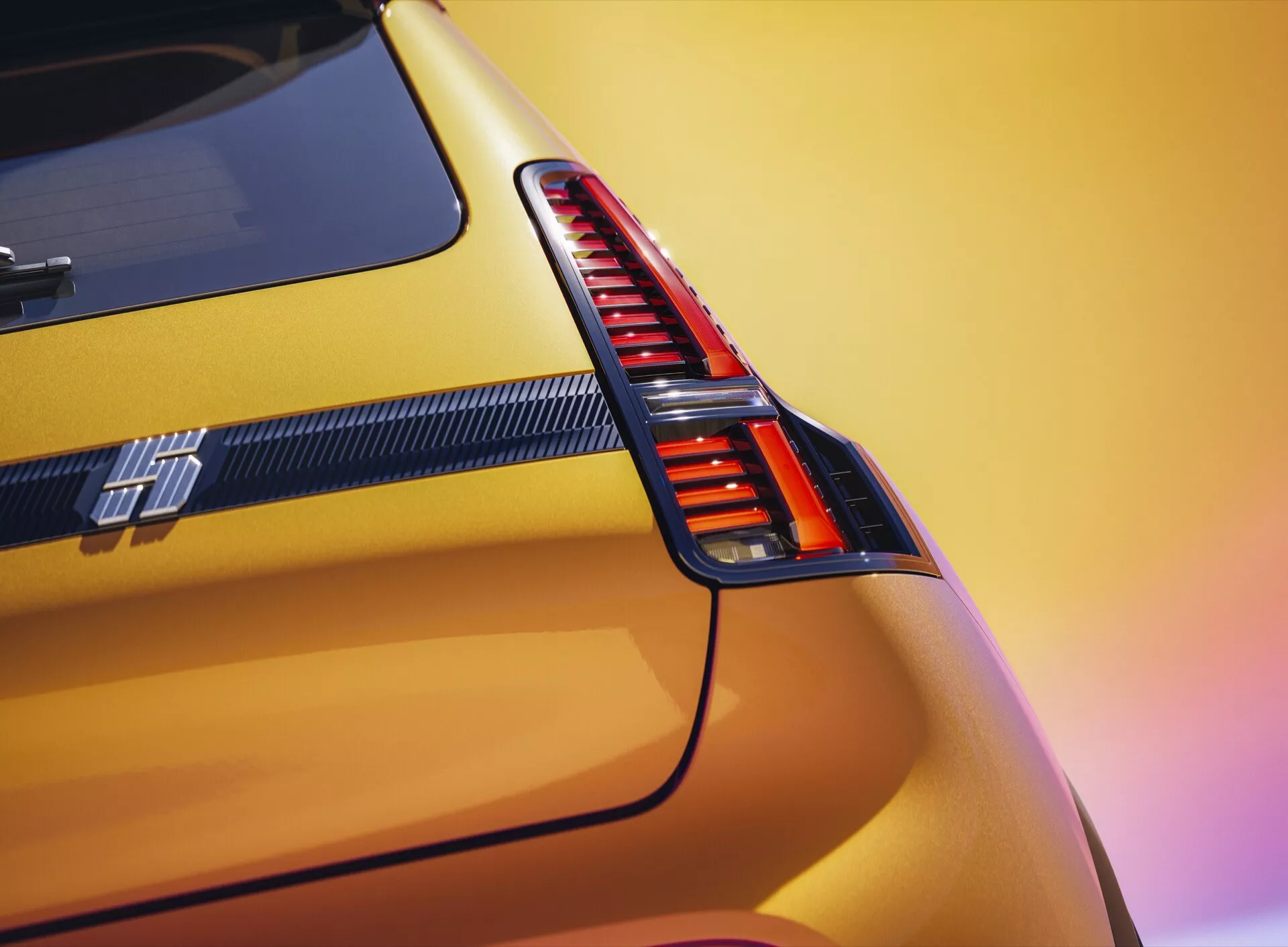 Detailní pohled na zadní část modelu Renault R5 2024 s vertikální červenou LED zadní světlou, živou kovově žlutou barvou a výrazným emblémem sportovní řady pod zadní světlou s prvky z uhlíkových vláken na pozadí rozmazaného západu slunce.