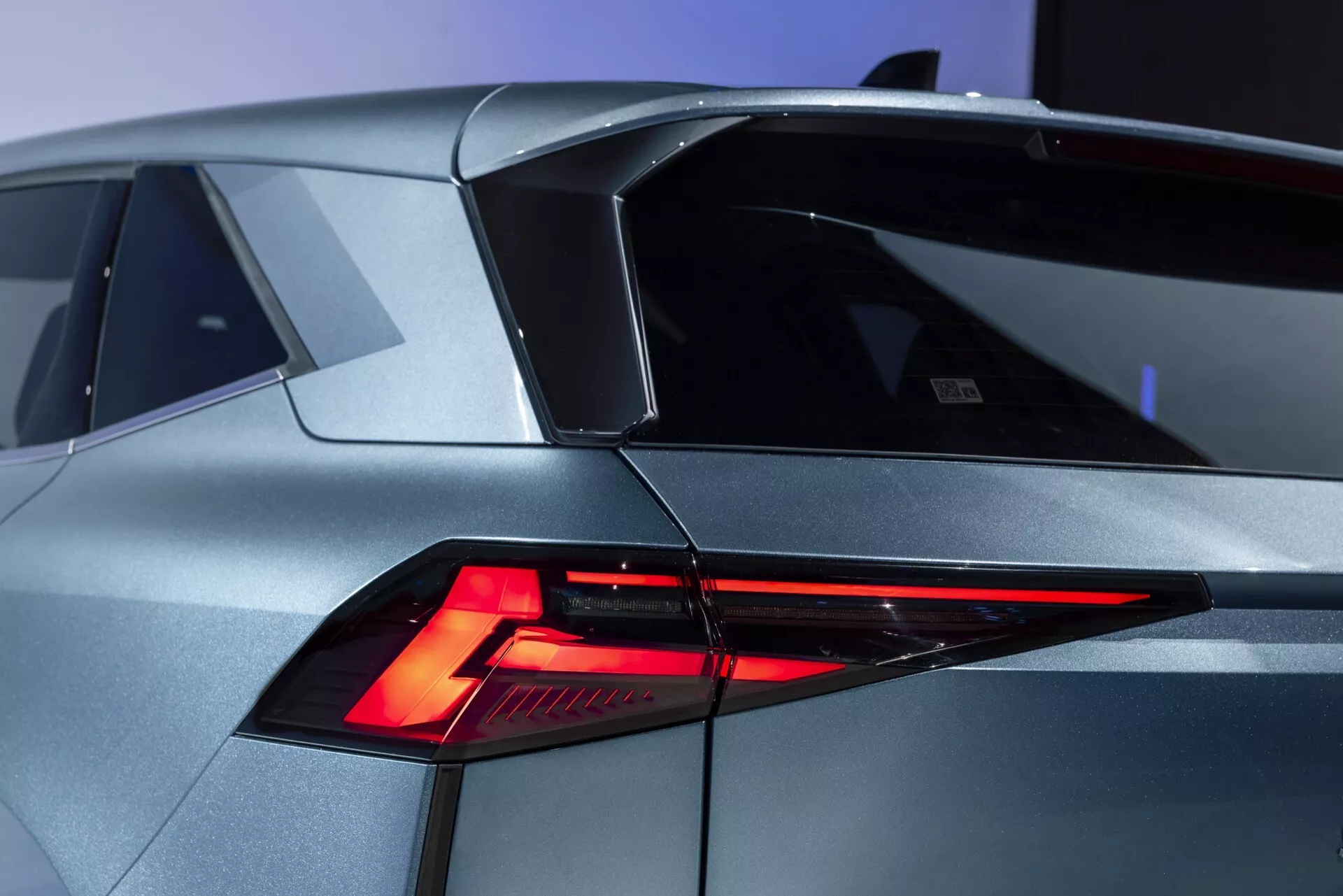 Detailní záběr moderního vozu Renault Symbioz 2024 (1. generace) z tříčtvrtečního zadního úhlu. Obraz výrazně zahrnuje ostrý, úhlový design LED zadních světel, skloněné zadní okno a výraznou linii střechy s malým spojlerem. Karoserie ukazuje futuristické geometrické tvary v kovovém stříbrném/šedém provedení. Na levé straně kufru je viditelné označení nebo znak, naznačující model nebo značku vozu. Další detaily zahrnují část připomínající víčko palivové nádrže na zadním levém čtvrtpanelu.