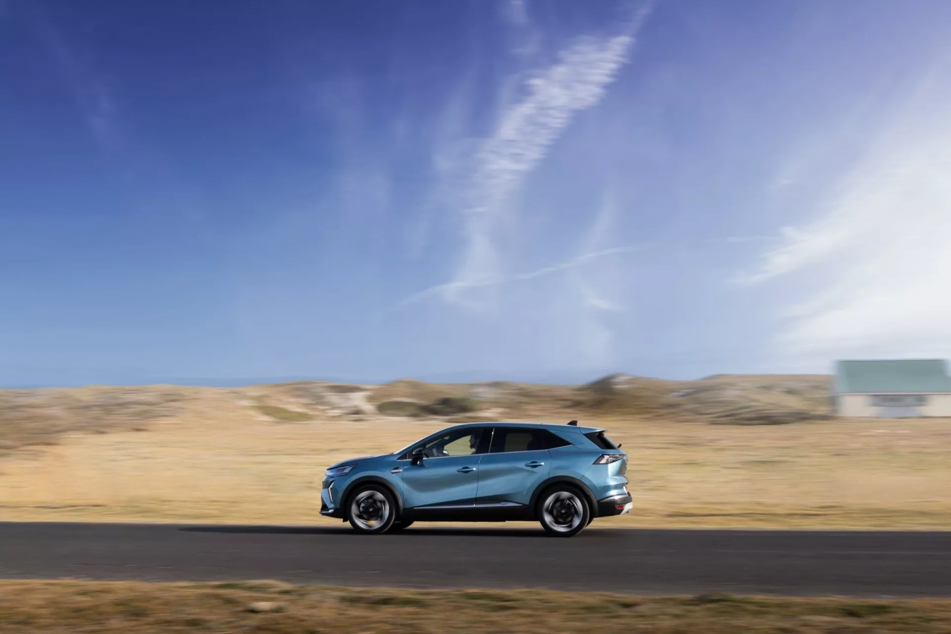 Dynamický boční profilový snímek pohybující se Renault Symbioz 2024 1. generace, pořízený technikou panning co na rozmazaném pozadí, který ukazuje jeho elegantní aerodynamický design, hliníková kola, boční zrcátka, integrované kliky dveří a kvalitní lak. Vozidlo je umístěno v dolní třetině rámečku proti modré obloze a pusté krajině, což zdůrazňuje rychlost, pohyb a schopnost automobilu v různorodém prostředí.