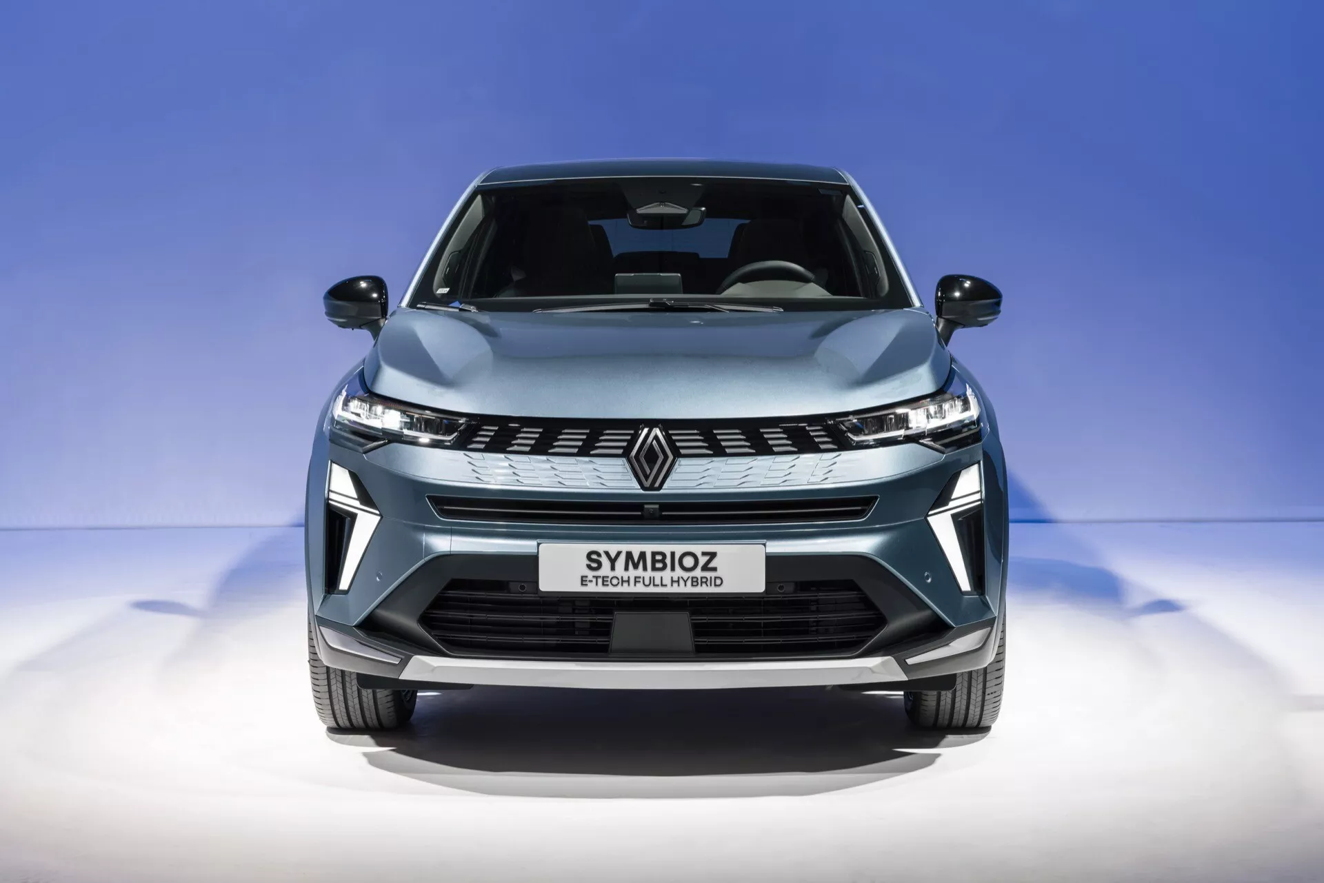 Blízký pohled zepředu na Renault Symbioz 2024 (1. generace) zachycený z mírného nízkého úhlu. Obrázek zdůrazňuje elegantní přední mřížku se stylizovaným znakem 'R', úhlové LED světlomety a aerodynamicky tvarovanou kapotu. Metalická světle modrá barva auta a jeho lesklý povrch jsou zvýrazněny proti čistému, přechodovému modrému pozadí. Text 'SYMBIOZ' na ploše pro registrační značku naznačuje moderní a možná futuristický design s prvky jako LED osvětlení, možnými autonomními systémy a pokročilou konektivitou nebo elektrickým pohonným ústrojím.