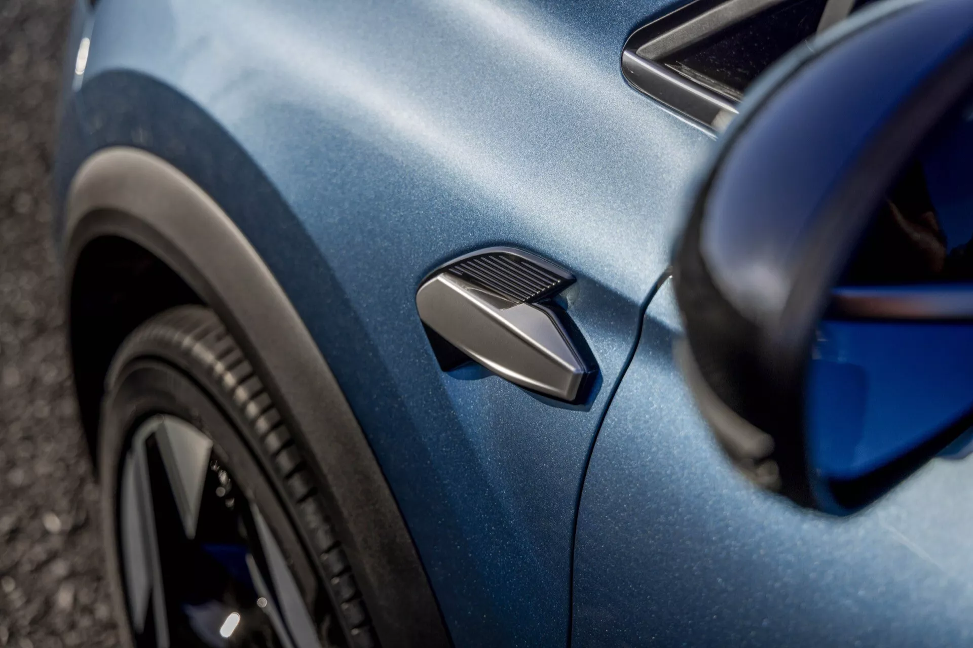 Detailní pohled na modrý Renault Symbioz 2024 (1. generace), zaměřený na zakřivení bočního panelu. Fotografie zvýrazňuje funkční postranní větrací otvor, část černého vícepaprskového předního kola a karoserii s kovovým povrchem. Mělký úhel a selektivní zaostření zdůrazňují aerodynamické a designové detaily vozu se zakaleným pozadím.