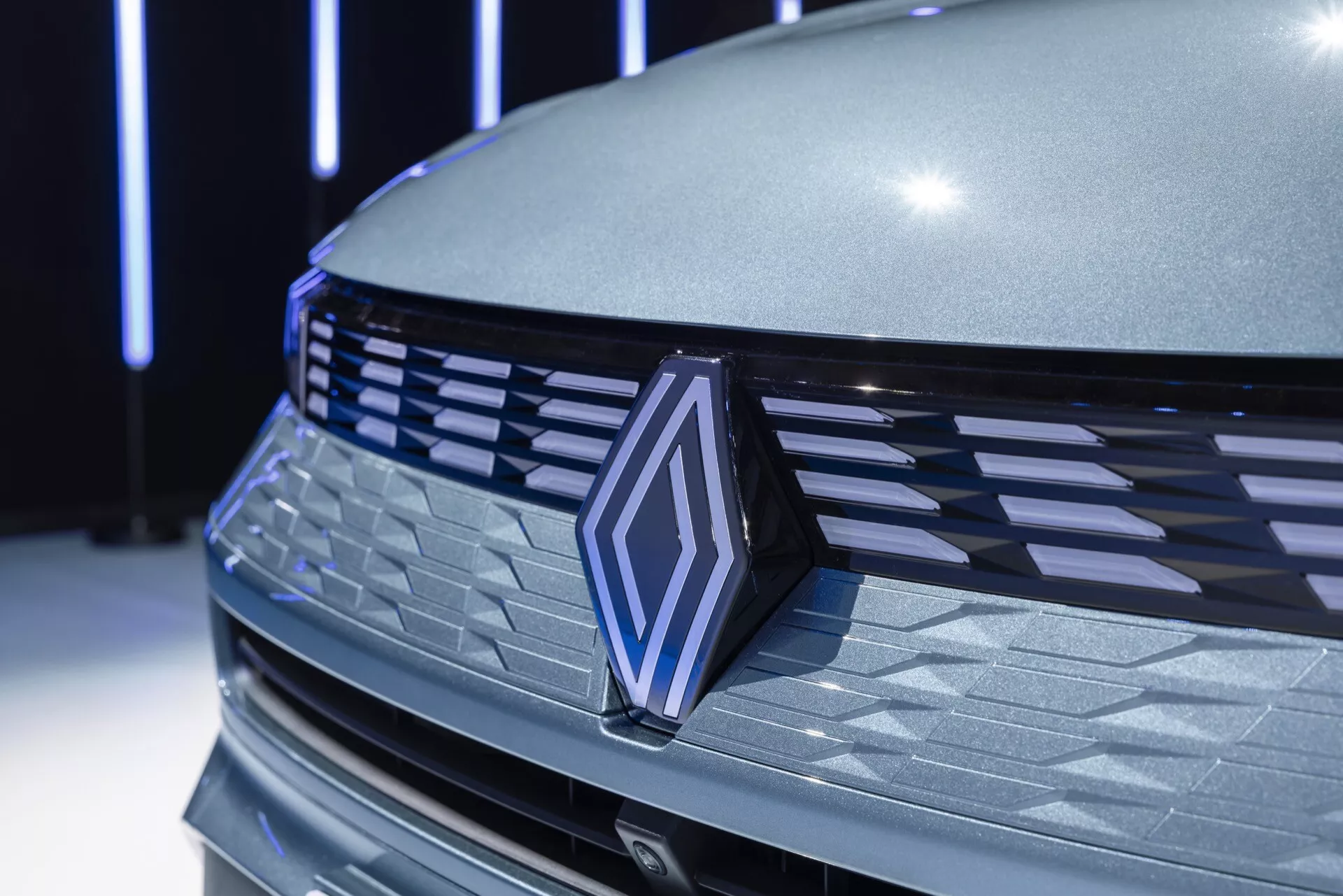 Detailní záběr přední části Renault Symbioz 2024 s důrazem na mřížkový vzor vodorovných lamel, výrazný emblém s chromovým rámečkem a tvarovanou kapotu. Kovový šedý povrch odráží měkké vnitřní osvětlení, čímž zdůrazňuje aerodynamický a luxusní vzhled vozu. Možné senzory pro asistenci řidiče mohou být skryté v symetrické mřížce, což naznačuje pokročilou technologii.