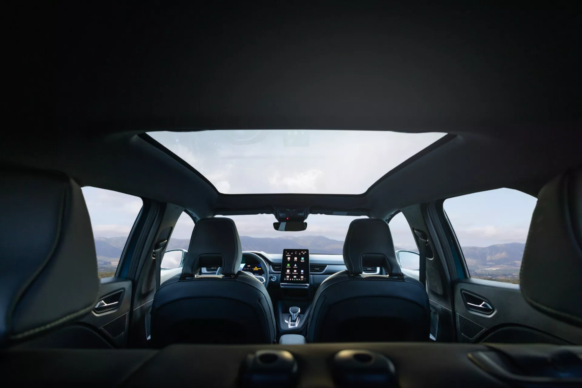 Záběr interiéru vozu Renault Symbioz 2024 (1. generace) z perspektivy zadního sedadla, který představuje moderní design s koženým čalouněním, výrazným prošíváním a pohodlnými konturovanými předními sedadly s opěrkami hlavy. Na středové palubní desce je výrazný vertikální dotykový infotainment systém, ovládací knoflíky klimatizace a digitální displej nebo HUD nahoře. Multifunkční volant je částečně viditelný. Pohled přes čelní sklo ukazuje horskou krajinu pod zamračenou oblohou, což naznačuje, že vozidlo stojí na malebném místě.