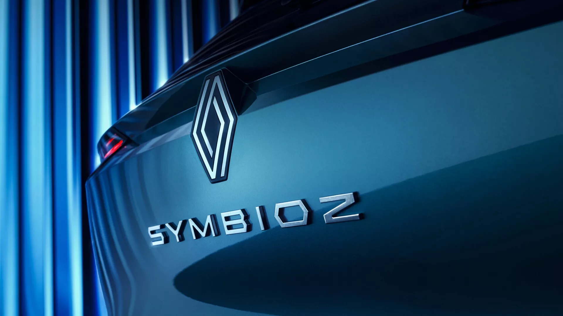 Detailní šikmý pohled na zadní část vozu Renault Symbioz 2024 (1. generace), zdůrazňující emblém značky a název modelu. Diamantový tvar emblému a slovo 'SYMBIOZ' ve velkých tiskacích písmenech bezpatkovým písmem jsou jasně viditelné na víku kufru, což naznačuje důraz na inovace a technologie. Měkký modrý odstín vrhá paralelní linie v pozadí, čímž vytváří elegantní a moderní atmosféru. Auto se jeví jako stojící, což zvýrazňuje jeho aerodynamické designové linie.