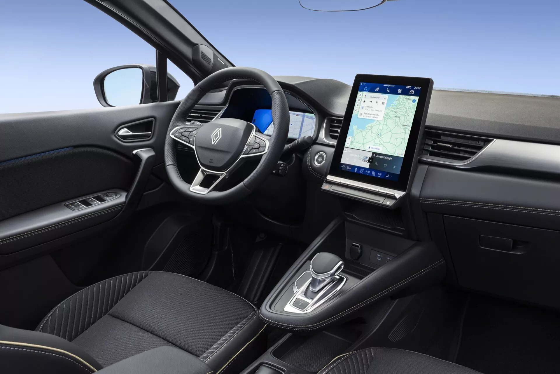 Interiér modelu Renault Symbioz 2024, zachycen z vyvýšeného, mírně pravého úhlu. Obsahuje multifunkční volant s emblémem výrobce, minimalistickou palubní desku naznačující digitální nebo head-up displej, velký vertikální dotykový infotainment systém zobrazující navigaci, moderní ovládání automatické převodovky, vysoce kvalitní černé sedadla s prémiovým čalouněním a dveře řidiče s ovládáním elektrického okna.
