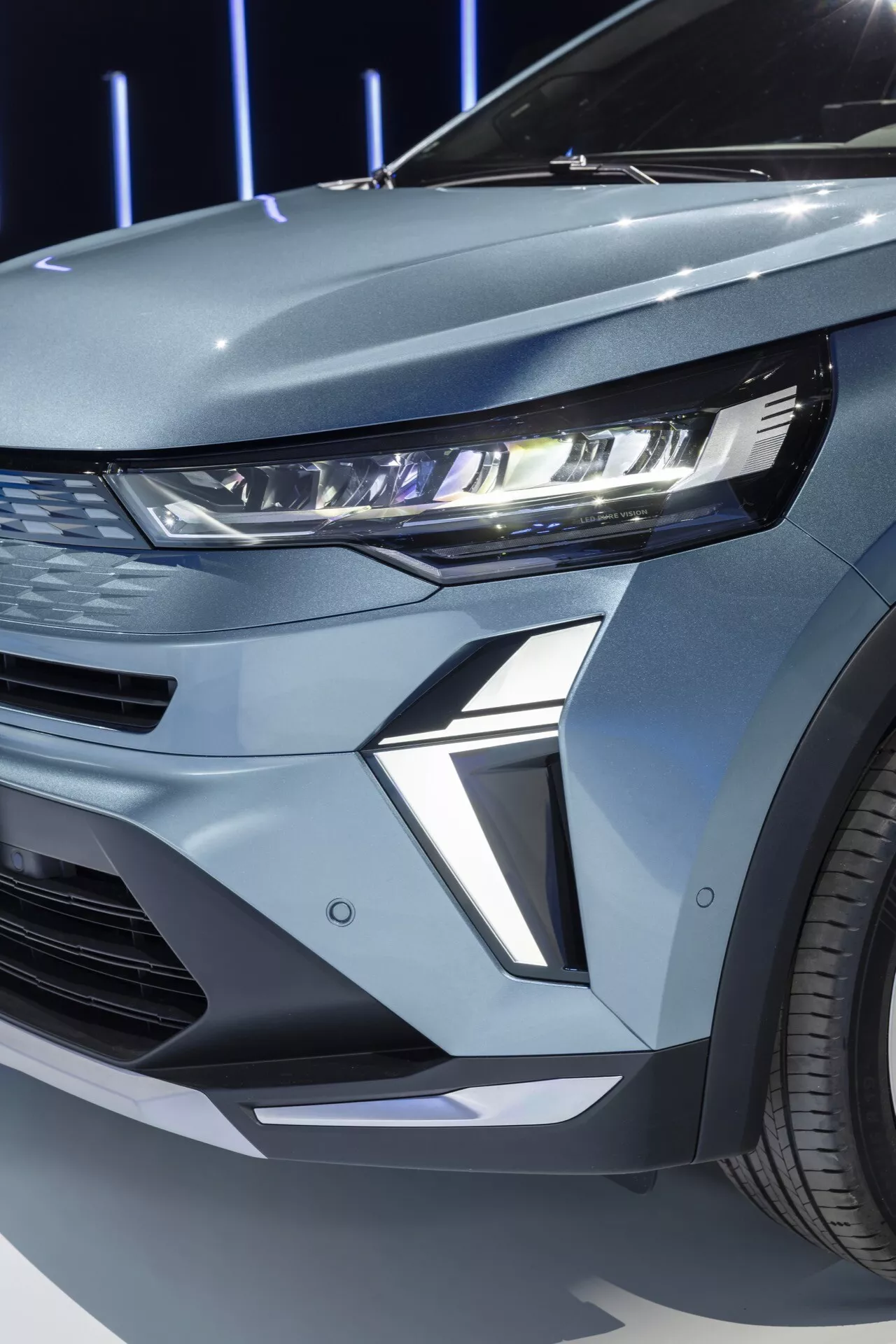Detailní pohled na přední levý kvadrant Renaultu Symbioz 2024 1. generace. Obrázek zachycuje moderní auto s elegantním LED světlometem s osvětleným denním svícením (DRL). Úzké světlometové pouzdro se táhne zpět do blatníku, což odráží současný automobilový design. Vysoce tvarovaný spodní část pod světlometem přidává vozu futuristický a agresivní vzhled. Viditelný segment přední mřížky naznačuje horizontální nasávání nebo dekorativní panel odpovídající sportovní estetice. Metalický povrch a viditelný mnohopaprskový kolo s litým ráfkem posilují prémiový vzhled.
