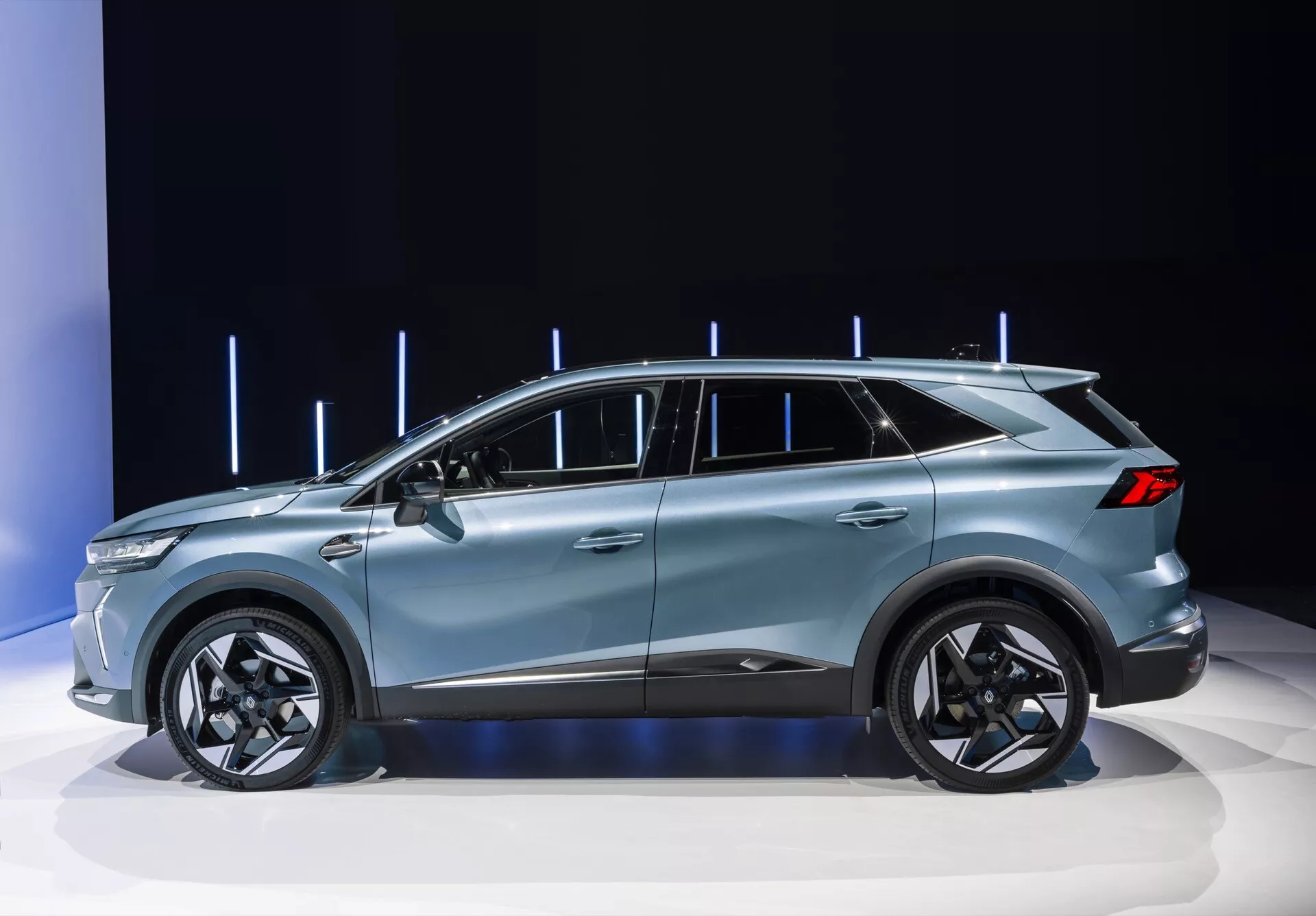 Obrázek zobrazuje Renault Symbioz 2024 (1. generace) na výstavě automobilů, kde je zřetelně viditelný elegantní boční profil moderního crossover SUV. Vozidlo se může pochlubit kupé podobnou siluetou, pěti dveřmi, velkými slitinovými koly a sofistikovanými designovými liniemi, vše zvýrazněné čistým, dobře osvětleným pozadím s vertikálními světelnými prvky. Design naznačuje prémiové funkce, jako jsou LED technologie, aerodynamická účinnost a možná pokročilé technologie asistence řidiče.