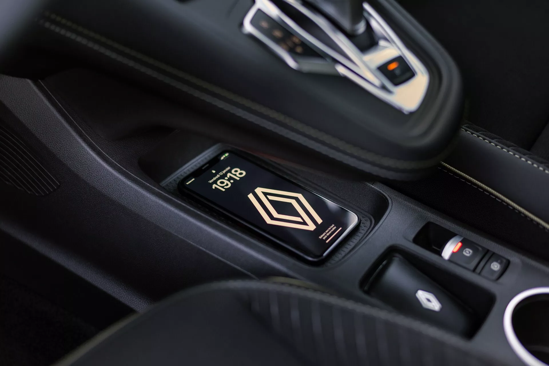 Pohled do interiéru Renault Symbioz 2024 na středovou konzolu s bezdrátovou nabíječkou, na které je telefon ukazující čas 10:18. Elegantní automatická převodovka s kovovými akcenty a ovládací panel v pozadí. Prémiový interiér s černou kůží a bílým prošitím.