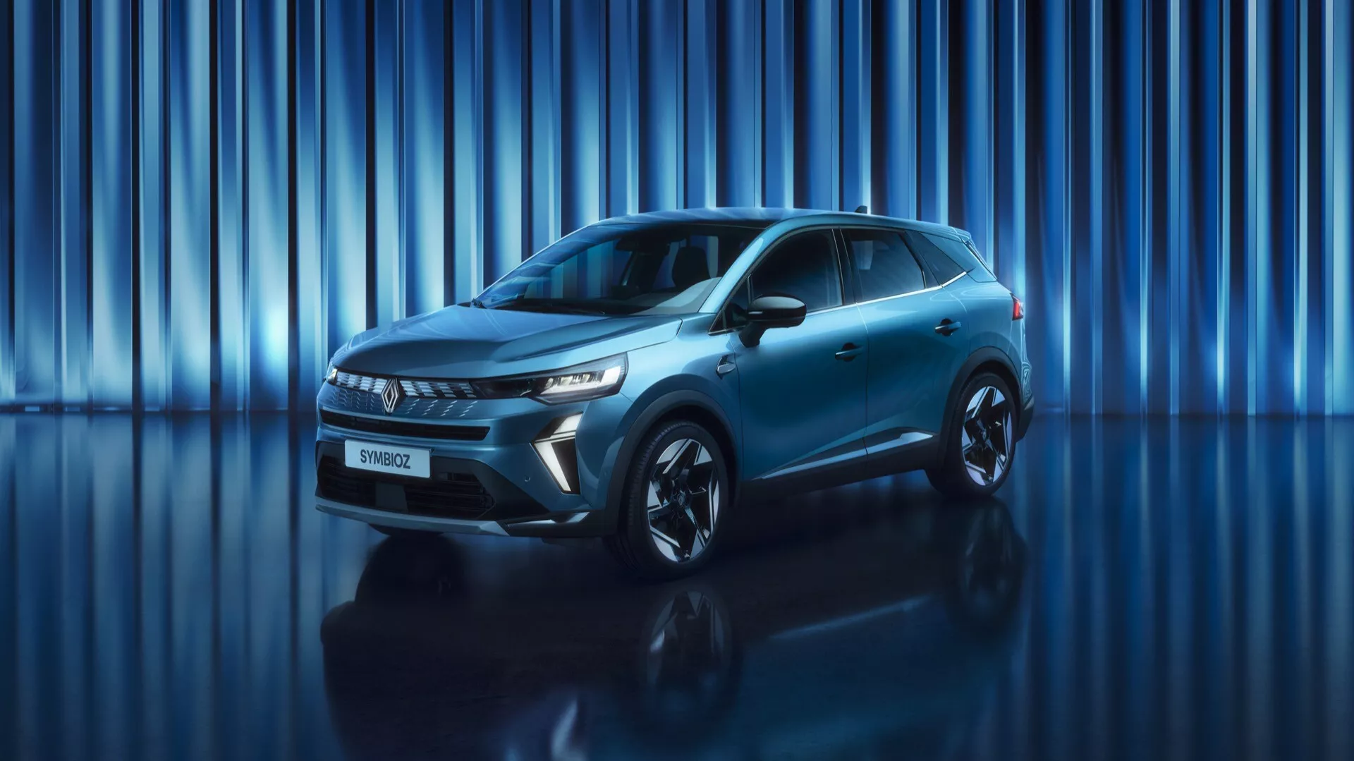 Obrázek zobrazuje SUV Renault Symbioz 2024 (1. generace) vystavené ve studiu s dynamickým modrým pozadím se svislými světelnými pruhy. Nízký až střední úhel ukazuje impozantní přední část vozu a elegantní boční profil. Vozidlo je celé na snímku, což zdůrazňuje jeho přítomnost a moderní design. Viditelné jsou ostré, úhlové LED světlomety a výrazná mřížka, spolu s hladkým předním nárazníkem, který může obsahovat bezpečnostní senzory. Boční profil odhaluje aerodynamické tvary, zapuštěné kliky dveří, aerodynamická zrcátka a velká kola z lehkých slitin. Metallic finished a sportovní postoj vozu jsou zvýrazněny studiovým osvětlením a odrazným povrchem pod ním.