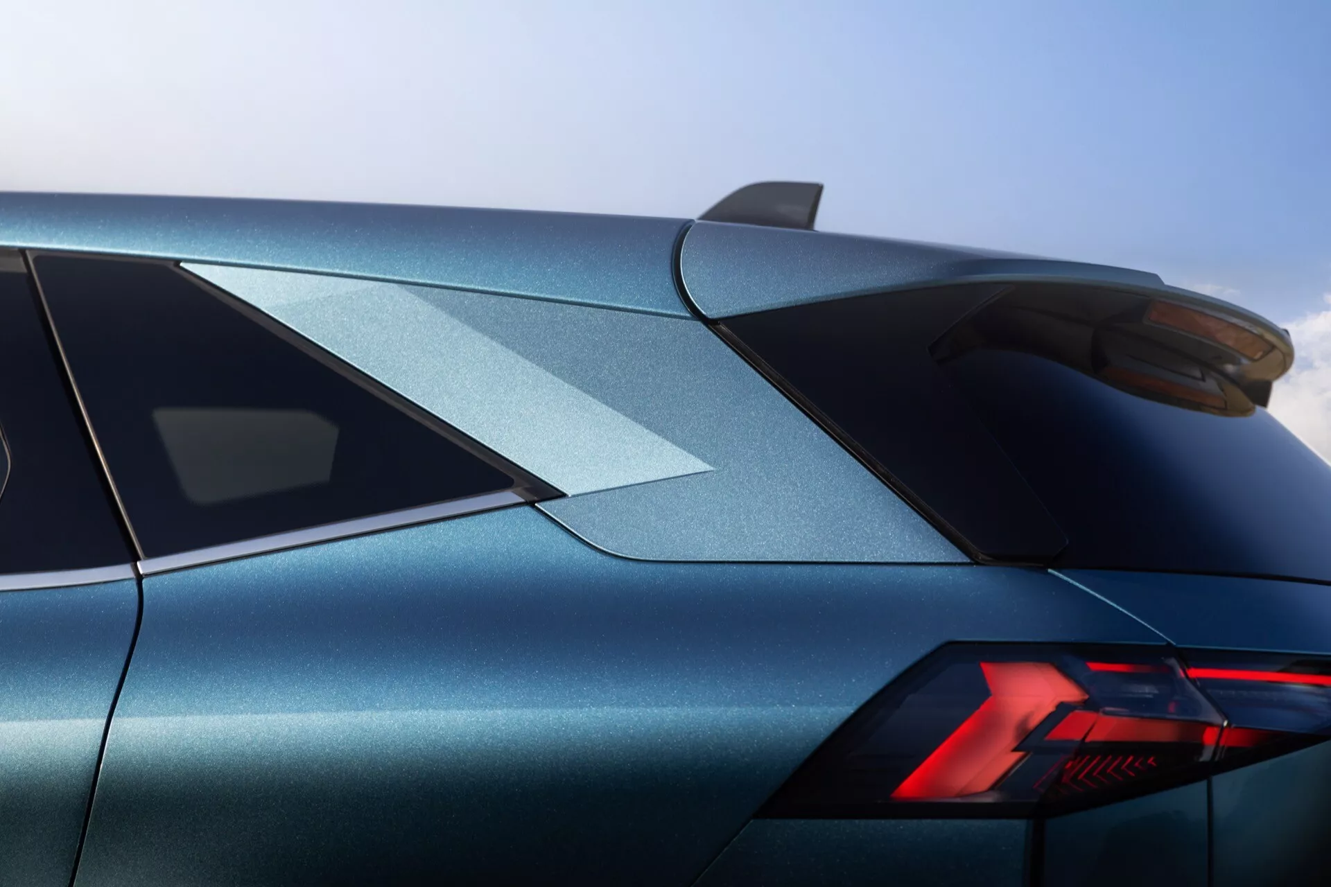 Tříčtvrteční záběr zadní části vozu Renault Symbioz 2024 z blízké vzdálenosti, zaměřený na horní zadní část. Obrázek ukazuje výrazně stylizované zadní světlo s moderními LED prvky ve tvaru 'Y'. Elegantní karoserie, dokončená v třpytivě metalické modrošedé barvě, obsahuje ostré, definované linie. Strmě skloněné zadní okno a namontovaný zadní spoiler podtrhují sportovní estetiku vozu. Na střeše je viditelná anténa ve tvaru žraločí ploutve, která dále indikuje moderní designové prvky. Vysoká kvalita zpracování a přesné detaily jsou z tohoto úhlu zřejmé, což naznačuje luxusní nebo výkonný vůz.