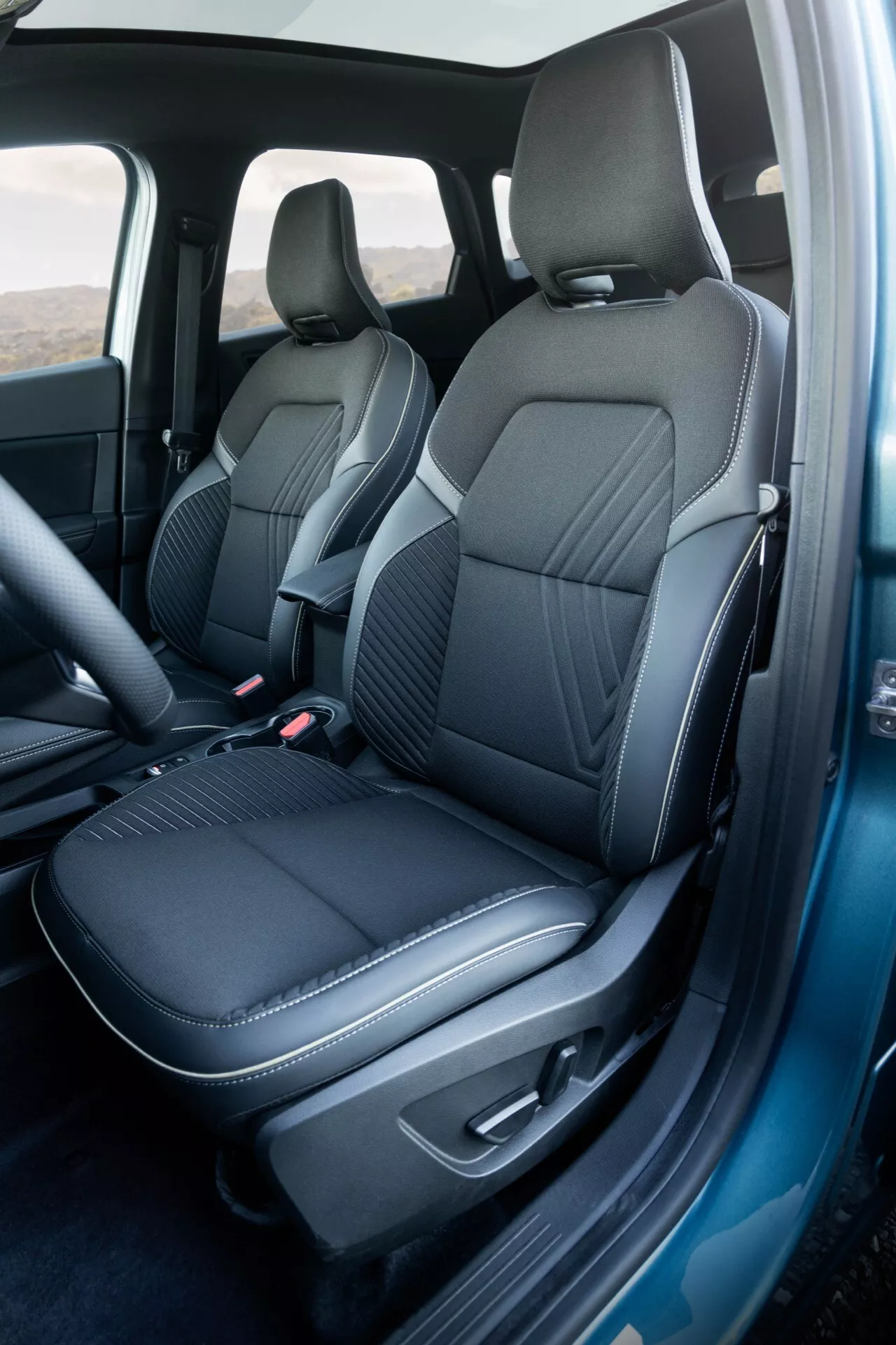 Detailní záběr na interiér modelu Renault Symbioz 2024 (1. generace), zaměřující se na přední sedadlo spolujezdce a část sedadla řidiče. Fotografie je pořízena z šikmého úhlu zvýhodňujícího stranu spolujezdce a ukazuje tmavé, prémiové čalounění s detailním prošíváním. Otevřené dveře u spolujezdce odhalují ergonomický design sedadel, hladké boční polštáře a centrální části s diamantovým prošitím, které zdůrazňují luxus a sportovnost. Integrované opěrky hlavy a minimalistický design bez viditelných nastavení potvrzují čistý, moderní vzhled. Panel otevřených dveří odpovídá materiálu sedadel s područkou a rozmazanými ovládacími prvky. Tmavý koberec ladí se sedadly. Viditelný B-sloupek a těsnění dveří naznačují péči o zvukovou izolaci, což ukazuje na vysokou kvalitu designu.