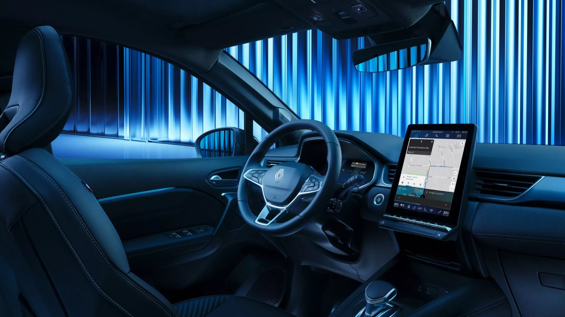 Pohled ze zadního sedadla na interiér Renaultu Symbioz 2024, zvýrazňující středově umístěný volant s integrovanými ovládacími tlačítky, digitální přístrojový panel a výraznou vertikálně orientovanou informační obrazovku zobrazující mapu. Kabina je osvětlena modrým ambientním osvětlením, což naznačuje moderní a technologicky orientovaný design, minimální fyzická tlačítka na středové konzoli a část dveřního panelu s integrovaným reproduktorem, což naznačuje prémiový zvukový systém.