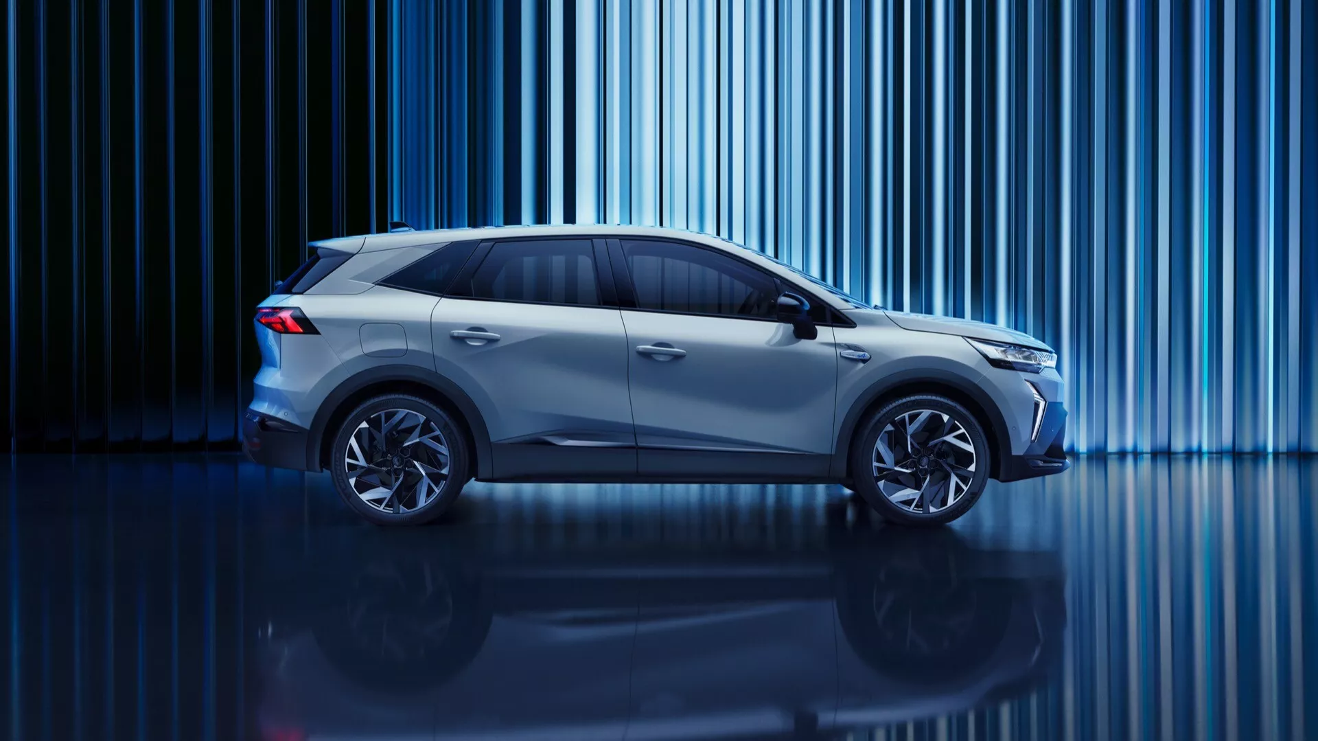 Snímka vo studiovém stylu Renault Symbioz 2024 (1. generace) z bočního profilu. Auto, kompaktní SUV nebo crossover se zvýšenou světlou výškou, je osvětleno kreativním osvětlením s modrým nádechem. Svislé světelné pruhy v pozadí a lesklá podlaha přidávají na technologické atmosféře. Viditelné jsou moderní designové prvky jako aerodynamické linie, velké ráfky kol a elegantní boční zrcátka. Tmavá boční okna naznačují pokročilé interiérové technologie a design napovídá energeticky úsporné inženýrství, možná elektrické nebo hybridní.