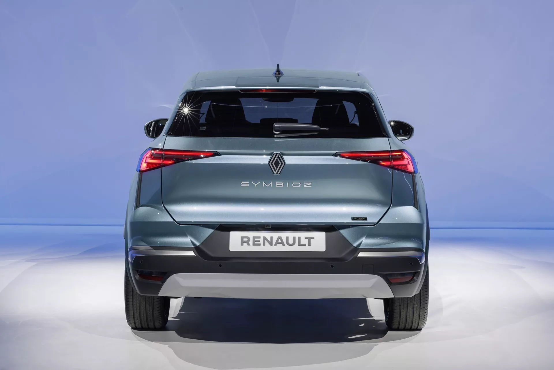 Pohled na záď Renault Symbioz 2024 ukazuje moderní designové prvky, včetně výrazných LED světel, prominentního loga značky, napsaného názvu a modelu, stěrače zadního skla, vysoko umístěného brzdového světla a zadních nárazníkových senzorů proti modrému gradientovému pozadí.