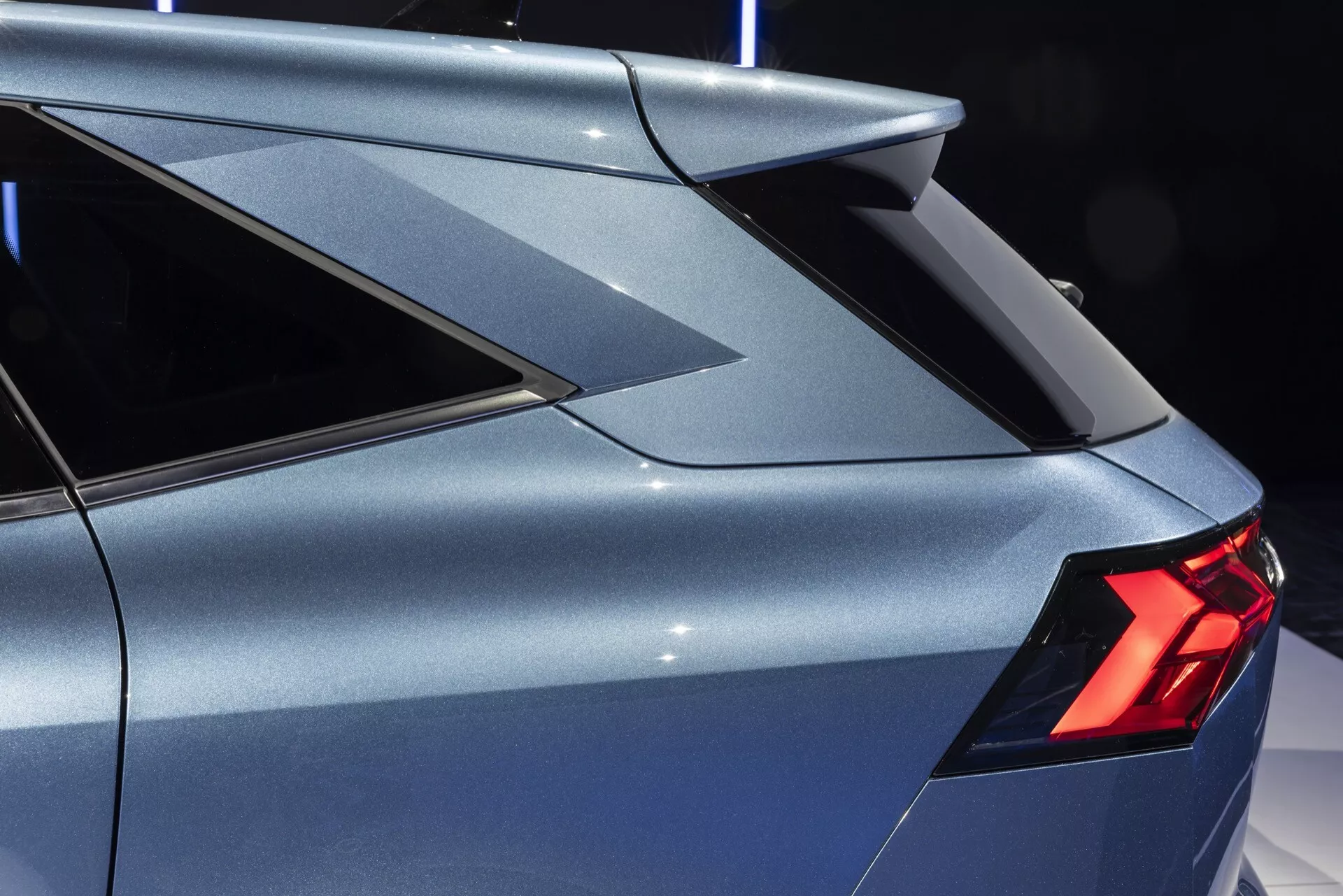 Detailní záběr zaměřený na zadní část Renaultu Symbioz 2024 (1. generace). Obrázek zachycuje vozidlo z mírně zvýšeného bočního úhlu, zdůrazňuje ostrý, hranatý C-sloupek s integrovaným sportovním zadním spoilerem. Viditelné jsou úzké, jasné LED zadní světla, stejně jako část zadních dveří a jemně zakřivená, aerodynamická karoserie. Metalická barva odráží osvětlení prostředí, což naznačuje jasně osvětlený profesionální prostor.