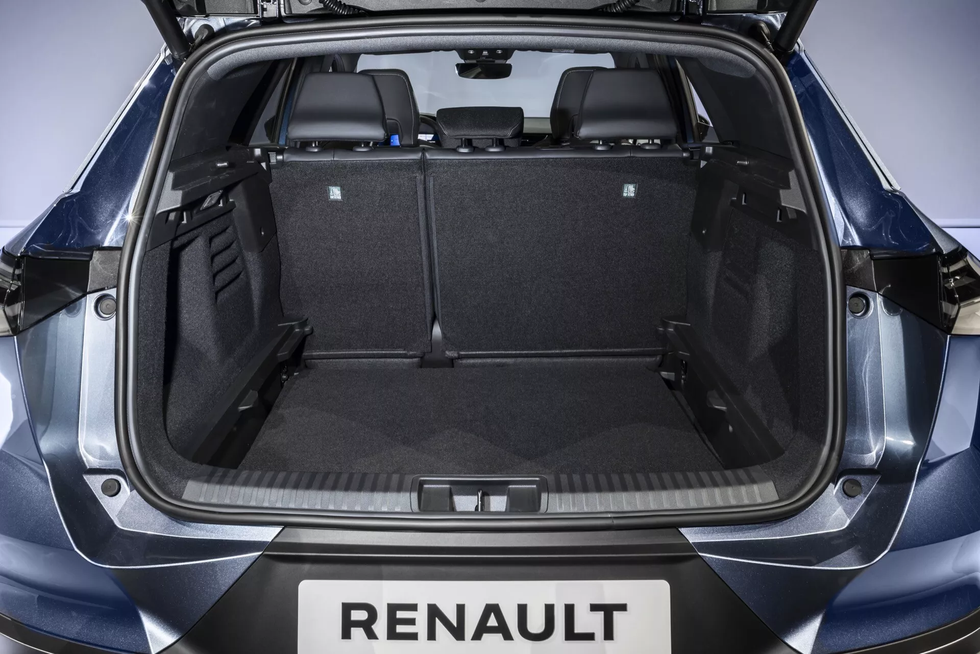 Vnitřní pohled na úložný prostor Renault Symbioz 2024 z otevřeného zadního poklopu, zobrazující symetrický prostor kufru s černým kobercem, plastovým obložením a sklopenými zadními sedadly pro rozšířené uskladnění. Viditelné branding zahrnuje 'RENAULT' pod poklopem.