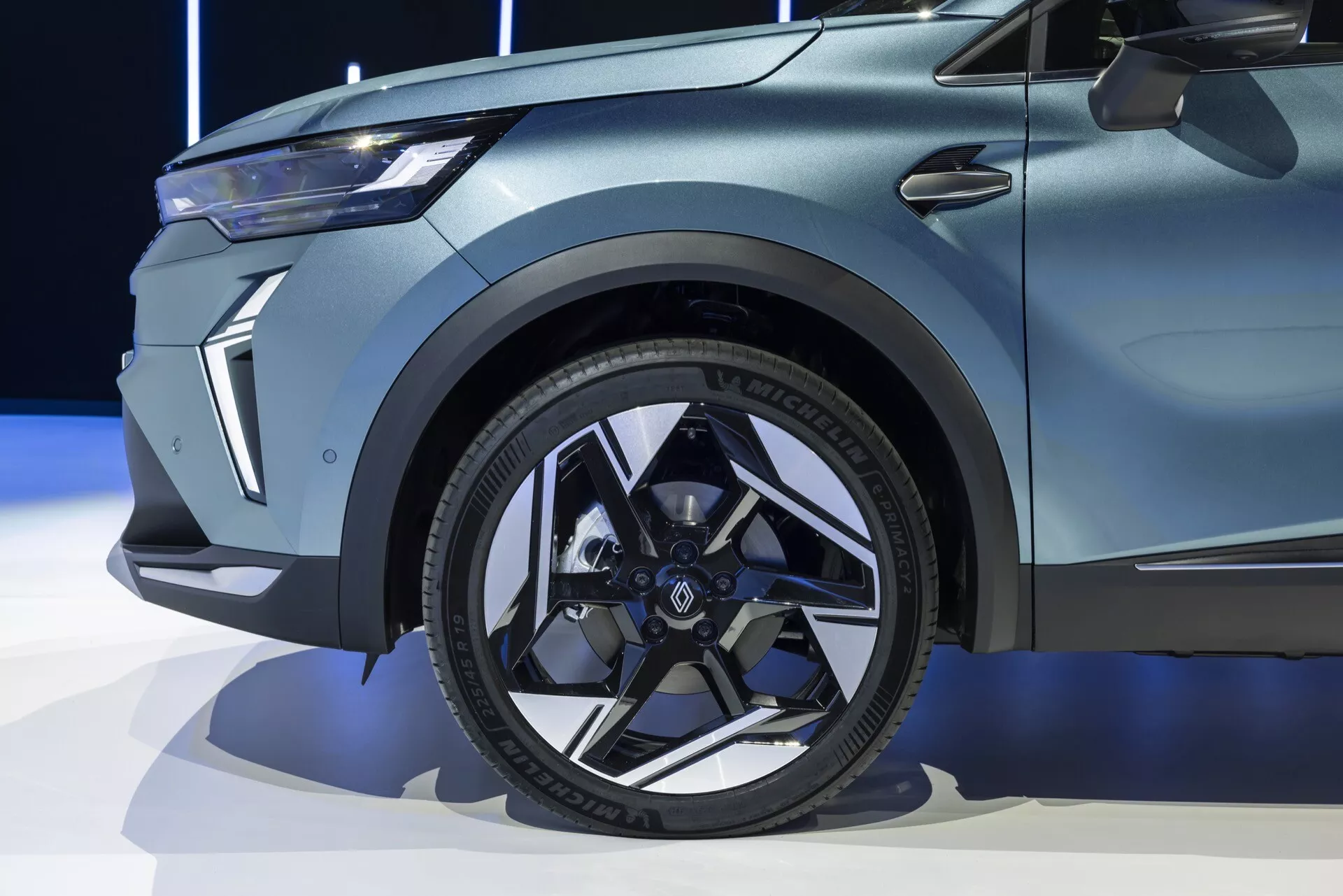 Detailní pohled na přední část Renaultu Symbioz 2024 (1. generace), zvýrazňující moderní černé hliníkové kolo s vícepaprskovým designem, nízkoprofilovou pneumatiku s výrazným nápisem na bočnici a ostré, svalnaté linie blatníku. Obrázek také zachycuje malou část dveří s výraznou linií a elegantní, úhlový design světel na přední fasádi. Lak vozu se zdá být matný nebo saténový, což naznačuje prémiový nebo speciální model vozidla.